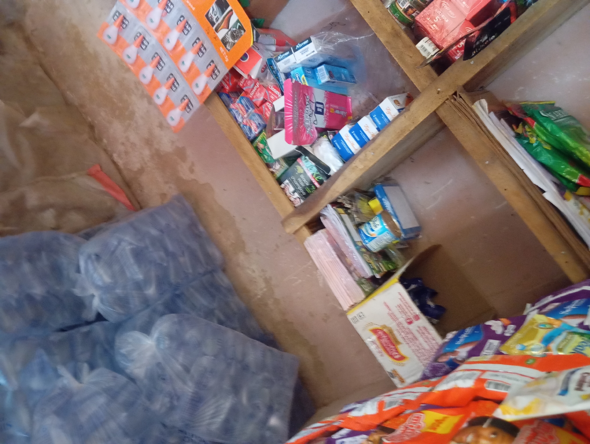 This image depicts the interior of a small shop in Jalingo, Nigeria. The shop is stocked with a variety of goods, including bottled water, packaged snacks, toiletries, and what appear to be small adhesive items. The shelving is constructed from wooden planks, some of which are precariously angled. In the foreground, large stacks of bottled water, wrapped in clear plastic, dominate the left side of the frame. In the background, shelves are filled with colorful packaging of various products. A close-up view of some of the products reveals brand names and imagery suggestive of everyday consumer items. The lighting appears to be ambient, casting shadows and highlighting the textures of the merchandise and the environment. The overall impression is one of a modest retail establishment in a local setting.