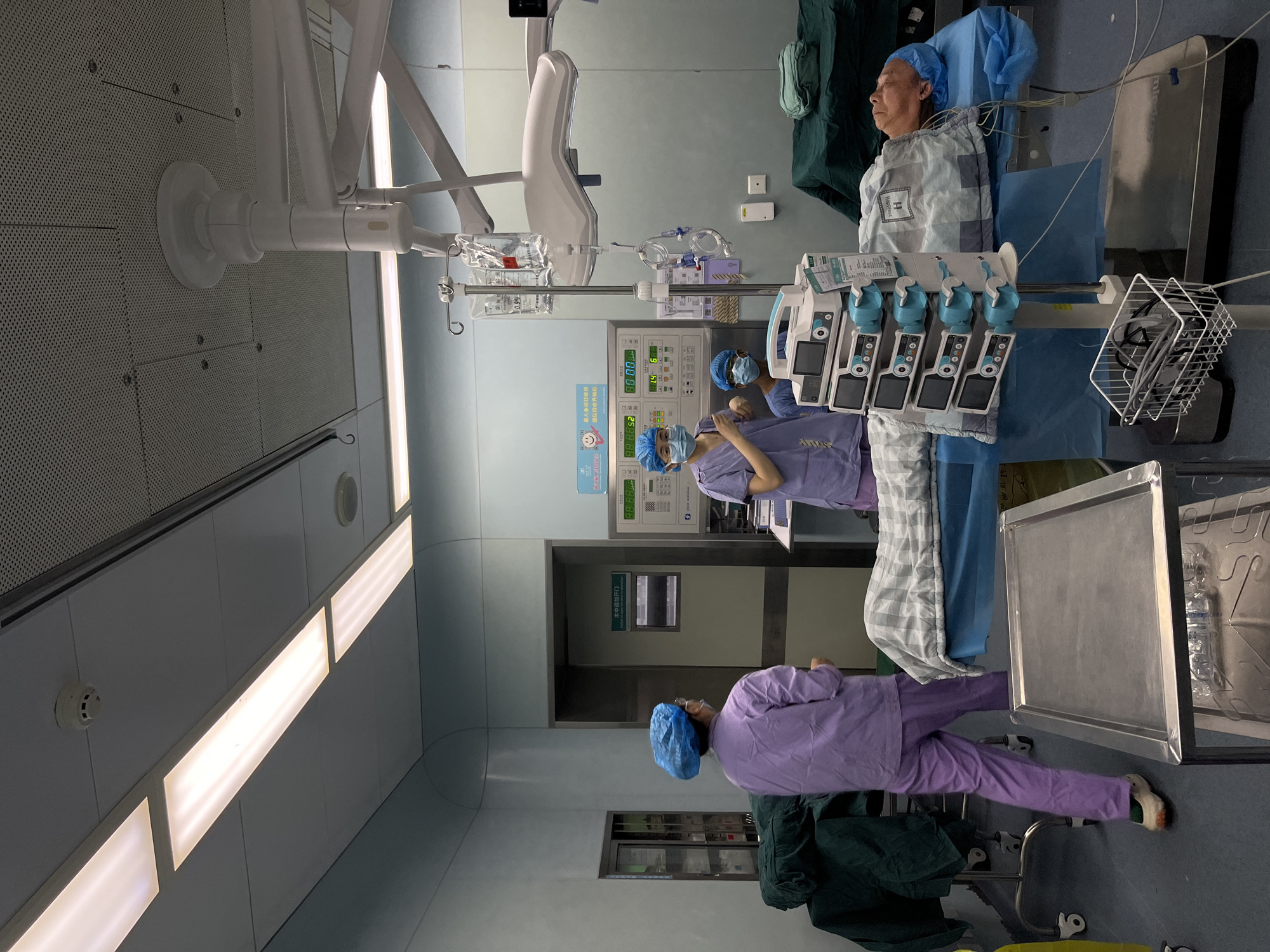 The image depicts an operating room scene, likely located in a hospital within the Yuzhong District of China. Two medical personnel are present, both wearing light purple scrubs, surgical caps, and masks, suggesting a sterile environment. One individual is positioned closer to a medical monitoring station and a patient lying on a surgical table. The other is further away, near a door with a sign written in Chinese which translates to "Do not open door during operation."
A patient is lying on the operating table, covered with a patterned blanket and surgical drapes. Various pieces of medical equipment are visible, including infusion pumps, monitoring equipment, an overhead surgical light, and a stainless steel tray. The room is clean and brightly lit with recessed ceiling lights. A window is visible near the door, suggesting the room is not entirely enclosed. Overall, the scene depicts a medical procedure in progress or preparations for one, inside a Chinese hospital.
