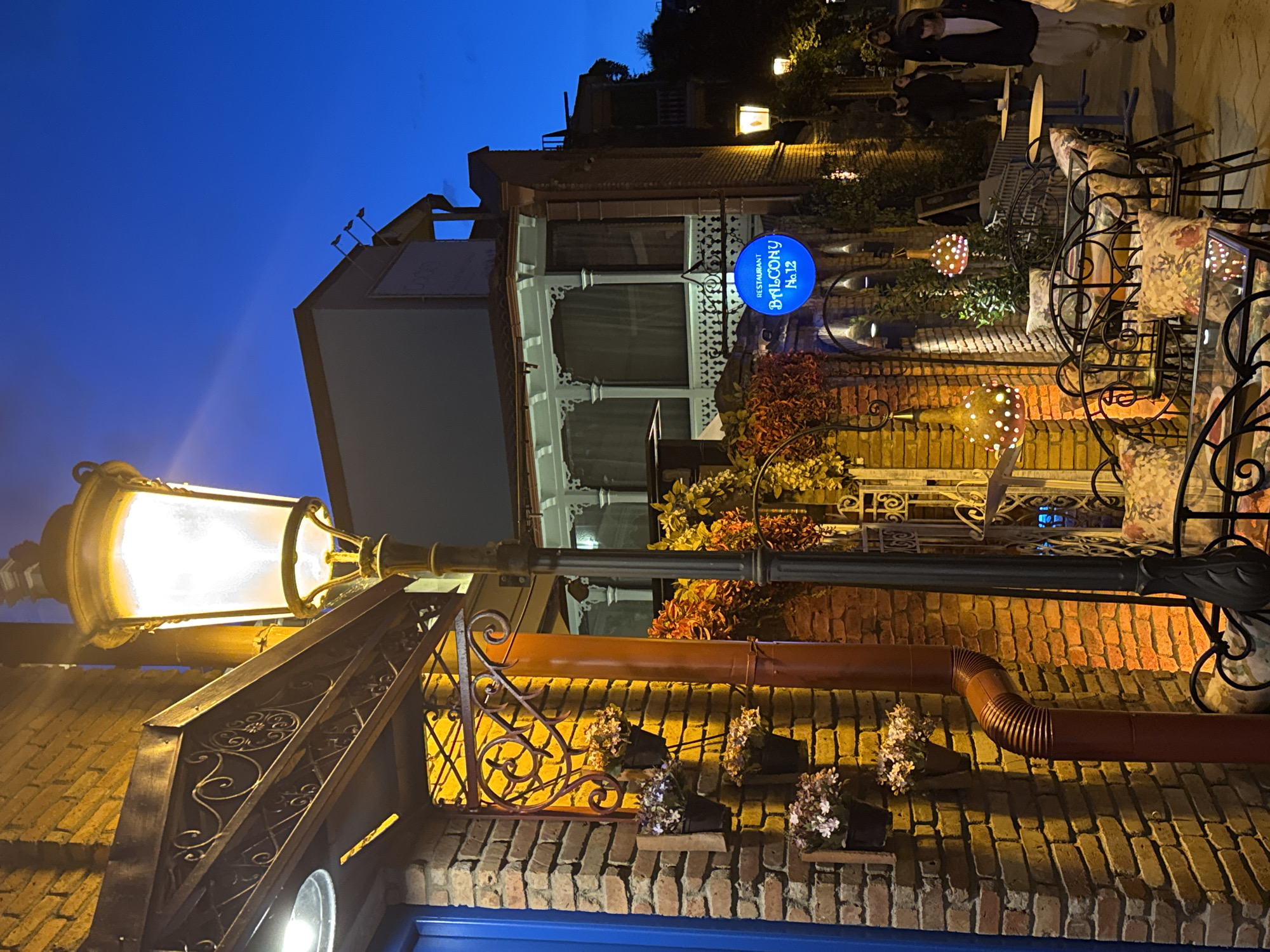 The image depicts an outdoor scene at night in Tbilisi, Georgia. A glowing street lamp dominates the foreground, casting light onto a brick wall adorned with flower boxes and an ornate wrought-iron structure. A brown pipe runs along the wall. In the background, a building with a white façade and a balcony is visible. A blue circular sign with the words "Restaurant Balcony No. 12" is attached to the building. Beyond the building, an outdoor seating area with wrought-iron chairs and tables is illuminated by string lights. Figures of people are blurred in the upper right corner. The sky is a deep blue, indicating dusk or night.
