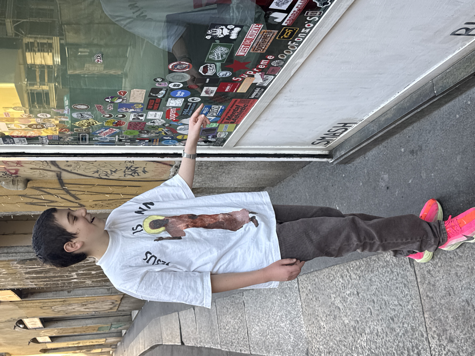 A person stands on a paved surface in front of a building in what appears to be an urban environment. The building has a glass surface covered in stickers, with the word "SMASH" visible on the lower right. The person is wearing a white t-shirt with an image of Jesus on it, brown pants, and bright pink running shoes. The person's arm is extended, pointing towards the stickers on the glass. Based on the OCR of the sticker on the window which reads "Ooofonso S" (a reference to the Instagram account) and the location of "SMASH" sticker, the image appears to be taken in the city of Milan, Italy, outside the "Slam Jam" store in Via Giovanni Lanza, 1.