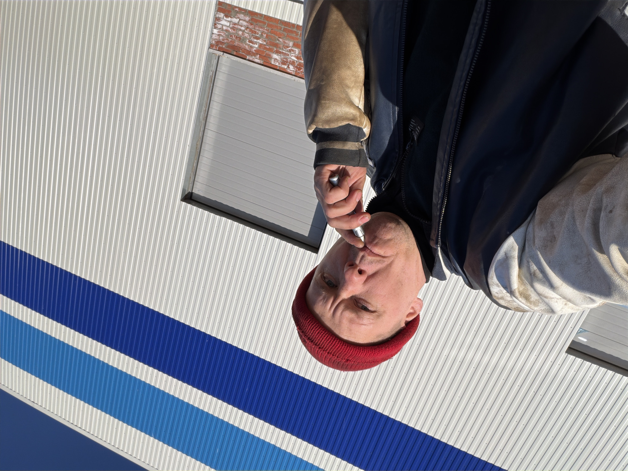 A medium shot of a man with a red beanie and a blue and tan jacket. He is holding a metal object to his mouth. The background is a white, corrugated metal wall with blue stripes. There is also a window with a red brick accent above it. The image appears to have been taken from a low angle and then rotated. It is not possible to determine the location as Chaikovsky, Russia from the given image.