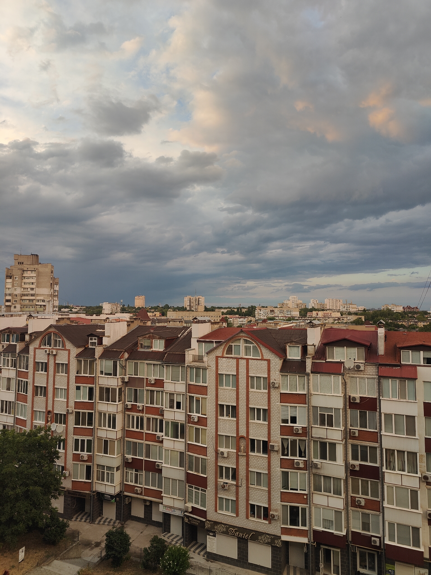 An exterior, slightly elevated shot captures a row of apartment buildings and other urban structures under a dramatic, cloudy sky in Chornomorsk, Ukraine. The foreground is dominated by the facades of several apartment blocks, characterized by their multi-story design, numerous windows, and balconies. These buildings display a combination of brick and white cladding, with red-tiled roofs and white window frames. Air conditioning units are visible mounted on many of the buildings. The lower levels of the buildings feature storefronts with signage, some of which are partially obscured.
To the left of the foreground apartment blocks, a large, lighter-colored building stands taller, also exhibiting a grid of windows. In the middle ground and background, a dense collection of city buildings extends towards the horizon. These structures vary in height and architectural style, with some appearing to be residential buildings and others possibly administrative or commercial. The overall impression is of a typical urban residential area.
The sky is a significant element of the scene, filled with heavy, gray clouds that suggest impending or ongoing rain. Patches of lighter sky and some warmer hues, possibly from the setting sun peeking through or reflecting off the clouds, are visible in the upper portion of the image. The lighting is diffused, characteristic of overcast conditions. The ground level visible in the lower portion of the image shows paved areas, walkways, and some vegetation, including a large tree on the left. The image lacks any visible people or discernible activity, presenting a static view of the urban environment.