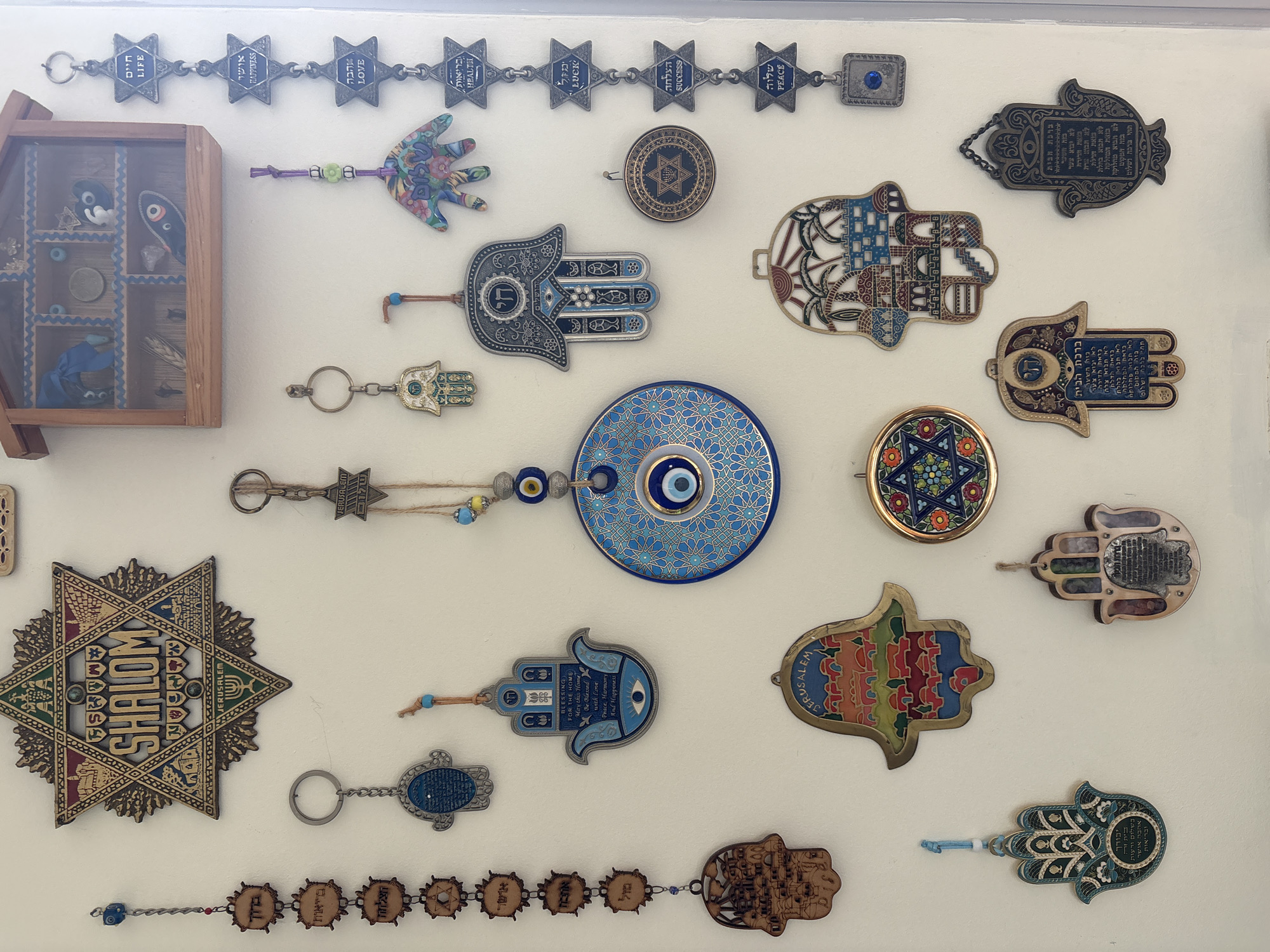 The image is a collection of Jewish-themed wall decorations. These include several Hamsa hand charms, some with Hebrew text or inscriptions, and some with evil eye symbols. There is also a Star of David decoration with the word "Shalom" (Peace) written on it, as well as a wooden shadow box with various small objects inside. Some charms have attached chains with Hebrew words such as "Life", "Happiness", "Love", "Health", "Luck", "Success", and "Peace". Other motifs include blessings for the home and the name Jerusalem. The background is a plain white wall. The image was not taken in Jesús del Monte, Mexico.