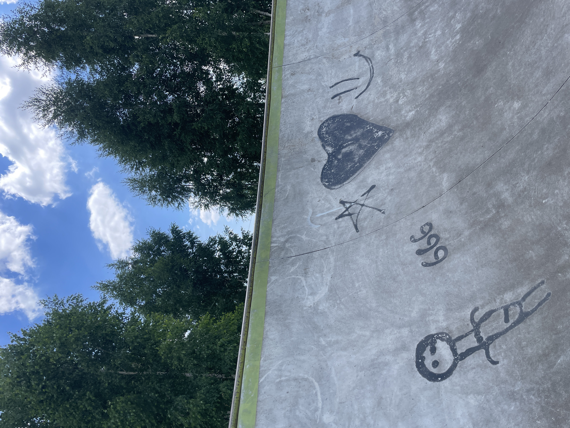 The image depicts a concrete skate ramp with graffiti art. The graffiti includes a stick figure, the number "666," a black heart with a star, and a smiley face. The skate ramp is outdoors, with trees and a blue sky with white clouds in the background. The location is identified as Laishevo, Russia.