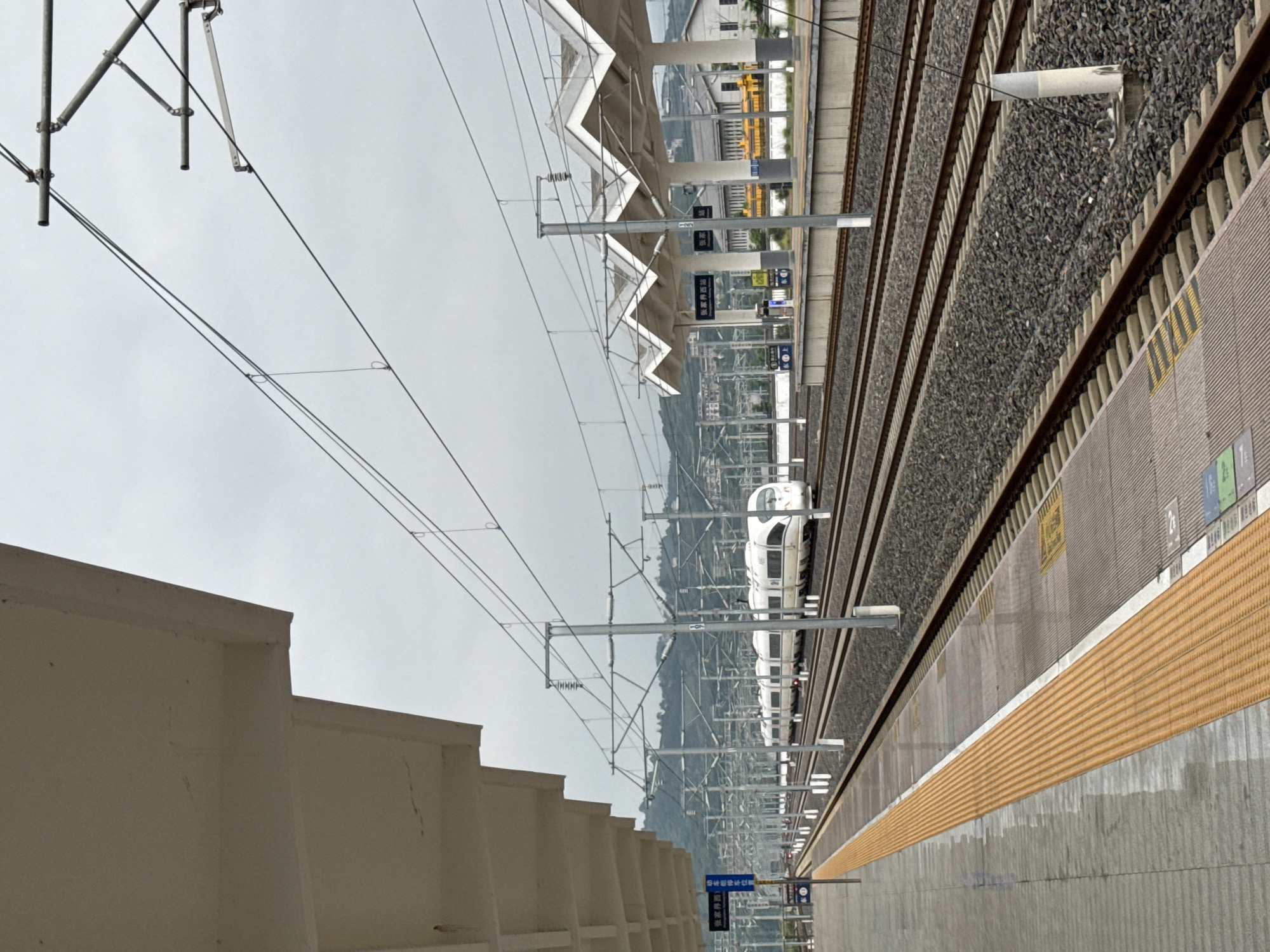 The image depicts a high-speed train at a station. A white train is visible on the tracks. The station platform is to the right with a yellow safety strip. Overhead power lines extend across the scene. A concrete wall borders the platform on the left. The sky is overcast. Signs in Chinese are visible on the platform. Based on these signs and information about the image, this is the Yongding District, China.