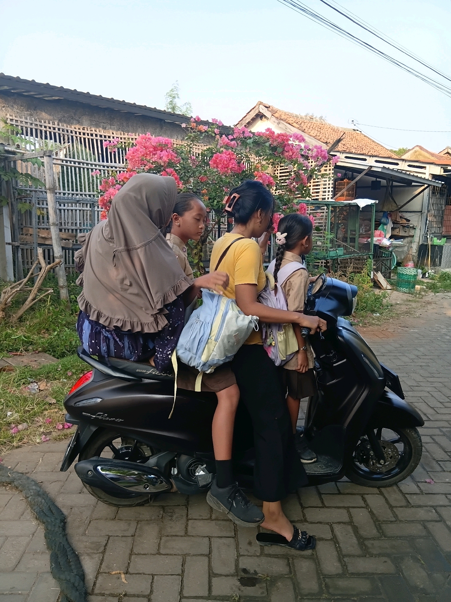 In the city of Cikal, Indonesia, four individuals are positioned on a dark-colored scooter. An adult female wearing a brown hijab and a patterned blue skirt is seated at the rear, with a young girl in a school uniform sitting in front of her, also wearing a hijab. A second young girl in a similar school uniform is seated in front of the adult, and an adult female wearing a yellow shirt and black pants is driving the scooter. Both young girls have backpacks. The scooter is on a paved brick surface, with a black curb and grass visible to the left. In the background, there is a house with a tiled roof and a fence adorned with pink bougainvillea flowers. The sky above is a pale blue.