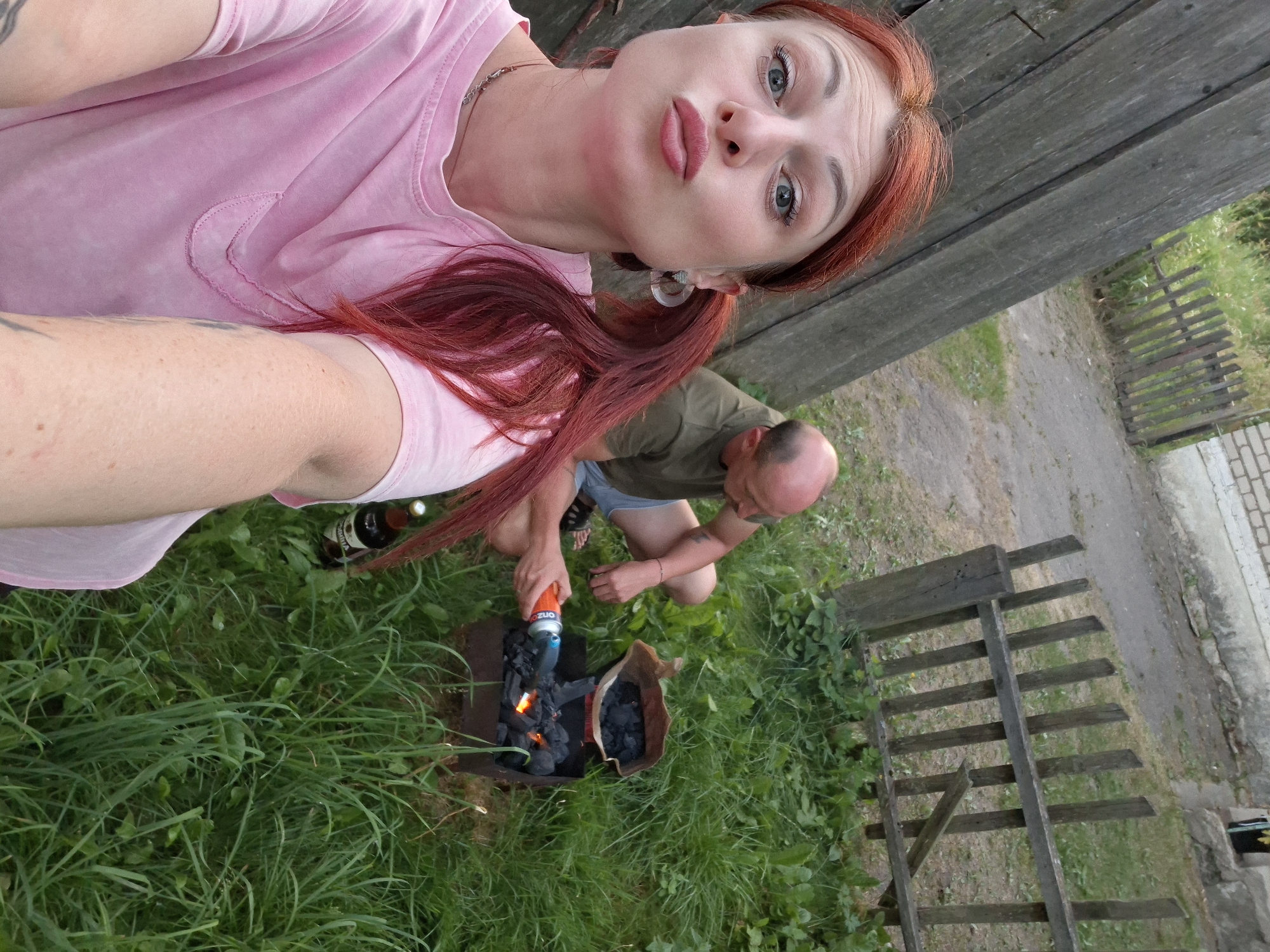 In this eye-level, slightly angled, outdoor photograph, a woman with striking red hair and makeup poses for a selfie while a man in the background crouches by a barbecue grill. The woman, wearing a pink t-shirt, looks directly at the camera with a playful pout, her long red hair styled in two pigtails. She has blue eyes and is wearing large, clear earrings.
The man, wearing a dark t-shirt and blue shorts, is focused on igniting charcoal in the grill. He is holding a can of lighter fluid, and flames are visible on the coals. Beside the grill is a paper bag filled with unlit charcoal. In the foreground, a bottle is visible lying on its side amidst lush green grass.
The scene appears to be in a rural or semi-rural setting, possibly a backyard, given the presence of a wooden fence and a paved path in the background. The lighting suggests it is daytime, likely late afternoon or early evening, with diffused light indicating overcast conditions or shade. The overall mood is casual and relaxed, hinting at a gathering for a barbecue. The backdrop includes a weathered wooden structure and more greenery. No discernible text is visible in the image.