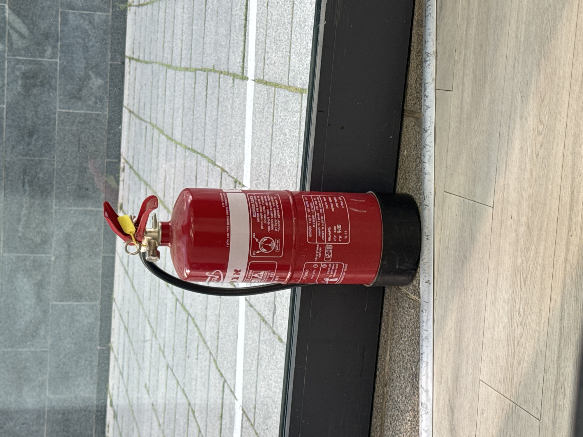 A red fire extinguisher lies horizontally on the ground. It is positioned between a light-grey, tile-covered walkway or sidewalk and a dark surface next to a light-colored wooden floor. The fire extinguisher features labels in Hebrew, including the word "אב" (father) and instructions, warnings, and weight information. The location is likely in Tel Aviv, Israel, given the Hebrew writing on the fire extinguisher.