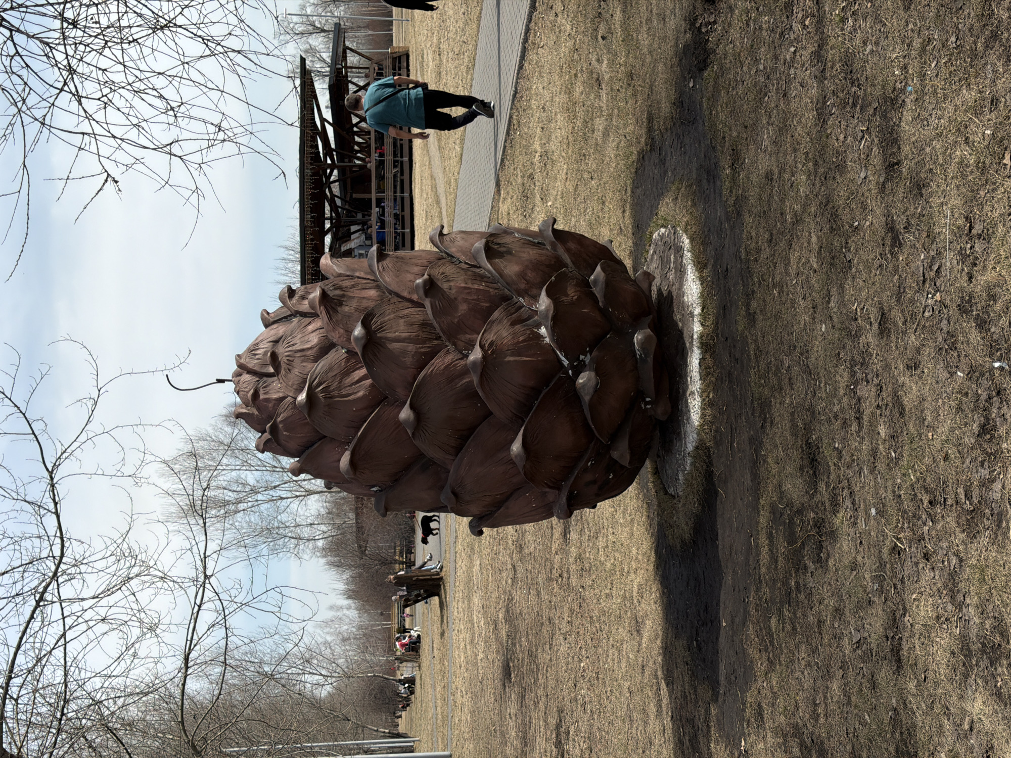 A large brown pine cone sculpture stands in a park, possibly in Krasnoyarsk, Russia. The sculpture rests on a circular patch of white-painted ground, surrounded by dry grass and bare trees. A person in a blue shirt and black pants walks in the background near a metal structure. Further back, people sit on benches. The sky is overcast.