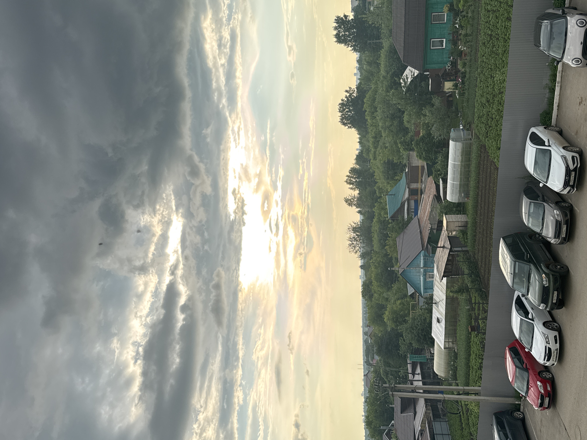 The image captures a view from a height, looking over a semi-rural area with a mix of residential buildings, gardens, and parking lots. The sky is dramatic, filled with heavy, gray clouds, but with shafts of bright sunlight breaking through, casting a warm glow on the landscape. Below the sky is a densely wooded area, with various shades of green foliage. Scattered within the trees are houses, some with traditional pitched roofs and others with more modern designs, including a bright green house with white trim. In the foreground, a row of parked cars is visible, including a red hatchback, a white sedan, a dark green SUV, and a silver compact car. A metal fence separates the parking area from a garden plot with greenhouses and neatly tilled rows. Utility poles and wires are also present, indicating infrastructure. The overall impression is of a tranquil, perhaps slightly overcast, day in a residential area on the outskirts of a town or village. Based on the provided location, this scene depicts a view within the village of Kondratovo, Russia.