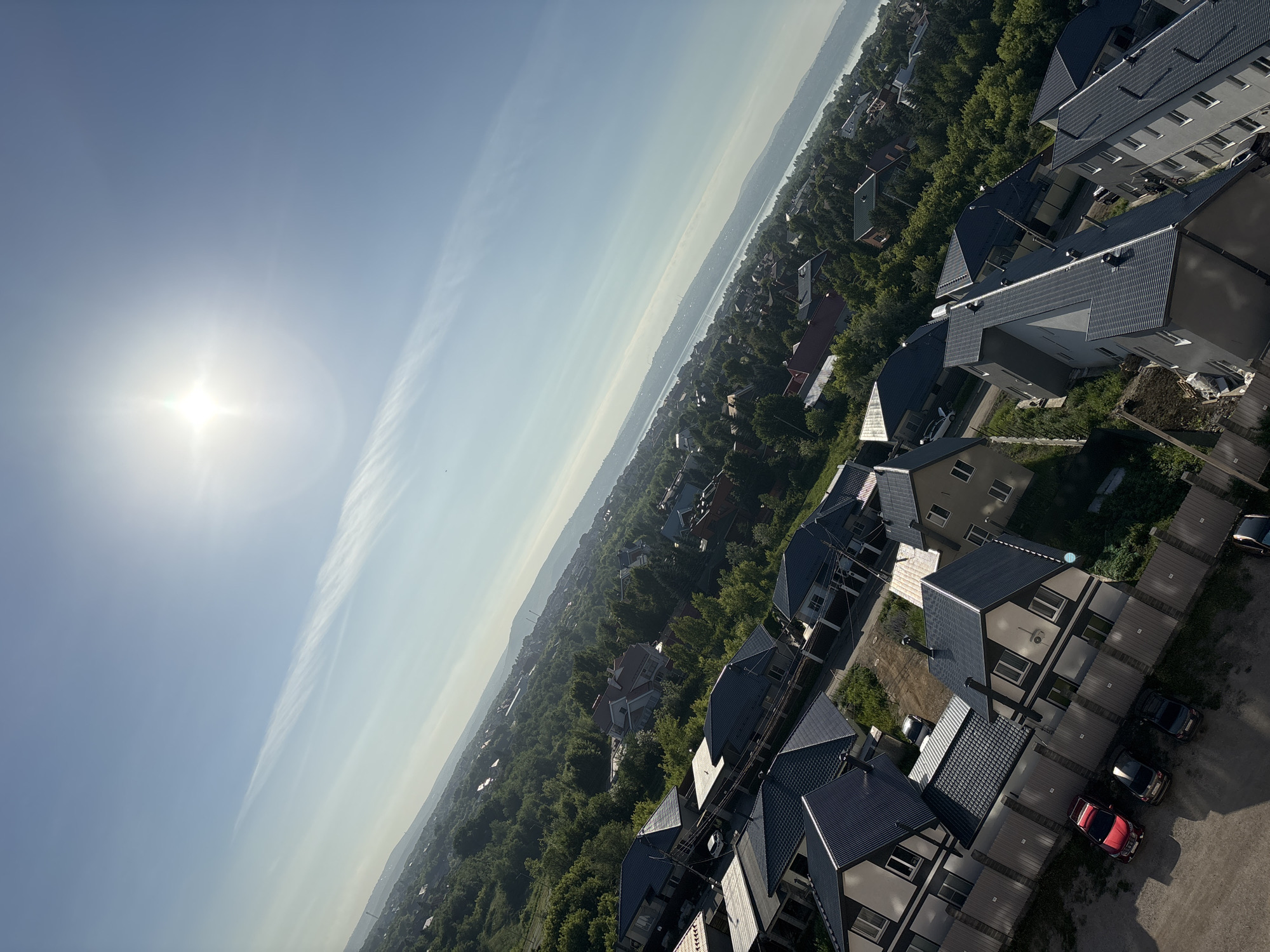The image is a high-angle, tilted aerial view of a suburban residential area under a bright, sunny sky with a few wispy clouds. The foreground is dominated by houses with dark gray pitched roofs, white or light-colored walls, and dark trim around windows. Some of these houses have fenced yards. Several parked cars are visible in a driveway and along a street, including a red SUV and two dark sedans. The houses are nestled among lush green trees, forming a dense treeline that extends into the midground. In the distance, a body of water, possibly a lake or river, is visible, bordered by hills or mountains on the far side. The overall impression is of a peaceful, well-developed residential area with abundant greenery and a clear, bright day.