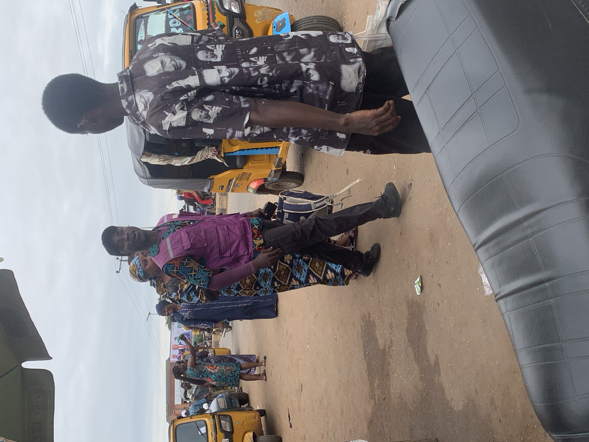 In Shagamu, Nigeria, under an overcast sky, a group of people are gathered on a dirt road near a yellow motorized tricycle. A man in a purple vest and black pants is speaking to another man wearing a black shirt with a repeating pattern of faces. To the left, a woman in a colorful dress is interacting with the group, holding a cell phone. Behind them, other individuals and a second tricycle are visible. The environment appears to be a marketplace or a busy street with shops in the background. The foreground is dominated by the textured seat of a motorcycle.