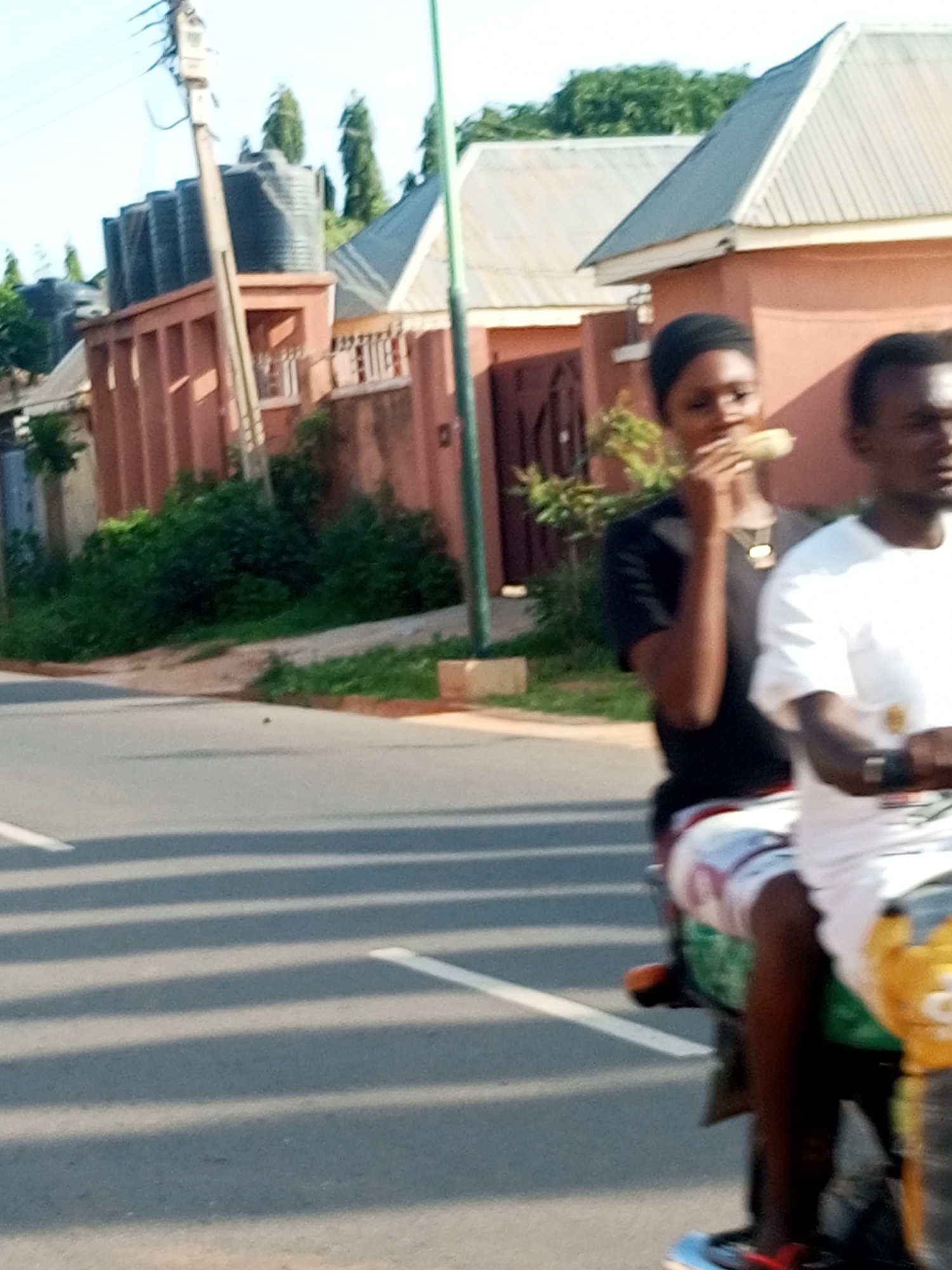 The image depicts a street scene in Tempure, Nigeria. In the foreground, two individuals are riding on a motorcycle. The person in the front, wearing a black headscarf and black t-shirt, is holding and eating a piece of corn. The person behind them, wearing a white t-shirt, is partially visible. The motorcycle appears to be green and orange. The road in the foreground has white painted lines and is casting shadows. In the background, there are buildings with terracotta-colored walls and grey roofs, along with lush green vegetation. Several large black water tanks are visible on top of one of the buildings. A utility pole with wires stands to the left. The overall lighting suggests it is daytime.