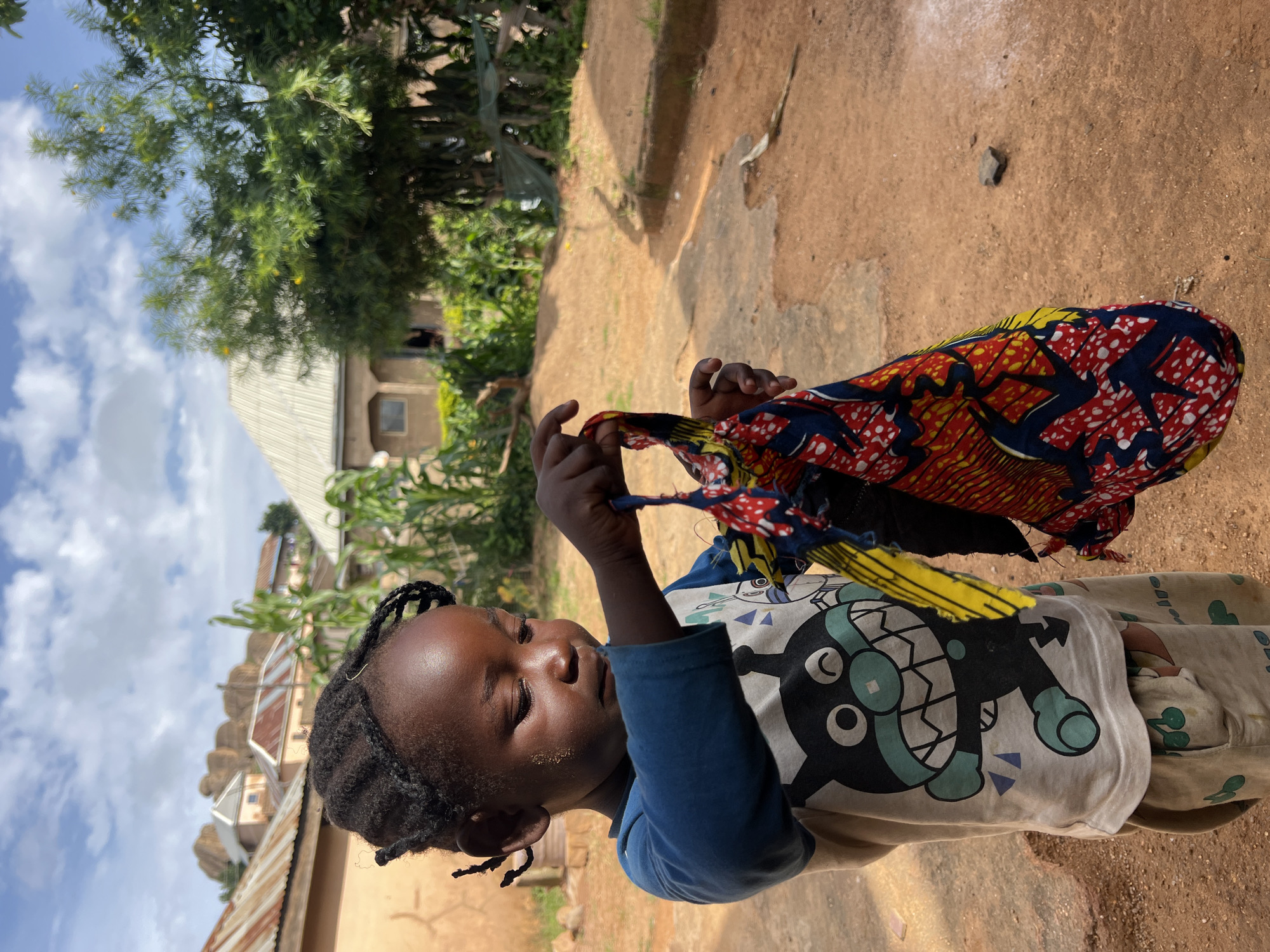 A young child with dark skin and braided hair stands in an outdoor setting, holding a colorful fabric in their hands. The child is wearing a light-colored t-shirt with a cartoon character printed on it and light-colored pants. The background shows a dusty ground, some vegetation, and buildings with corrugated metal roofs, suggesting a rural or semi-urban environment consistent with parts of Jos, Nigeria. The sky is partly cloudy. The child is looking down at the fabric, which has a pattern of red, yellow, and blue. The overall lighting suggests it is daytime.