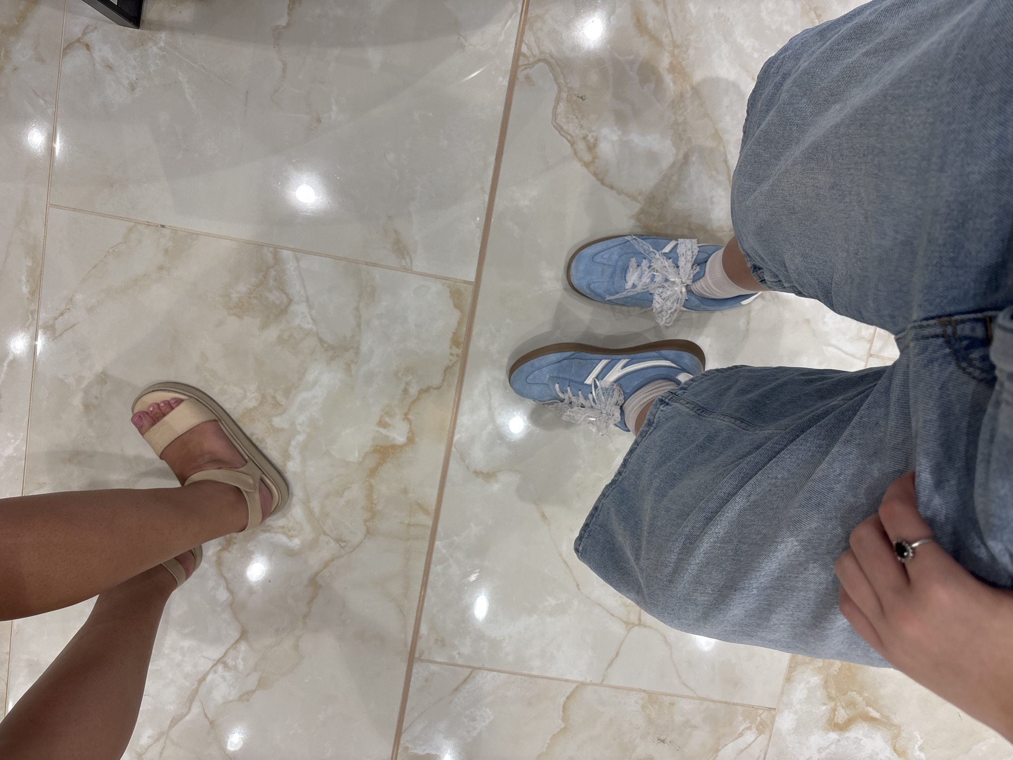 The image shows a close-up of two pairs of legs and feet standing on a marble-tiled floor. One person is wearing light-colored sandals and the other is wearing blue sneakers with white laces. Both are wearing casual clothing, one with light blue denim jeans and the other partially visible in darker trousers. A hand with a dark stone ring is visible in the foreground resting on the jeans. The floor is made of large, polished marble tiles with natural veining patterns and reflections of light. The location is not specified in the image, therefore it cannot be determined if it is in Vratsa, Bulgaria.