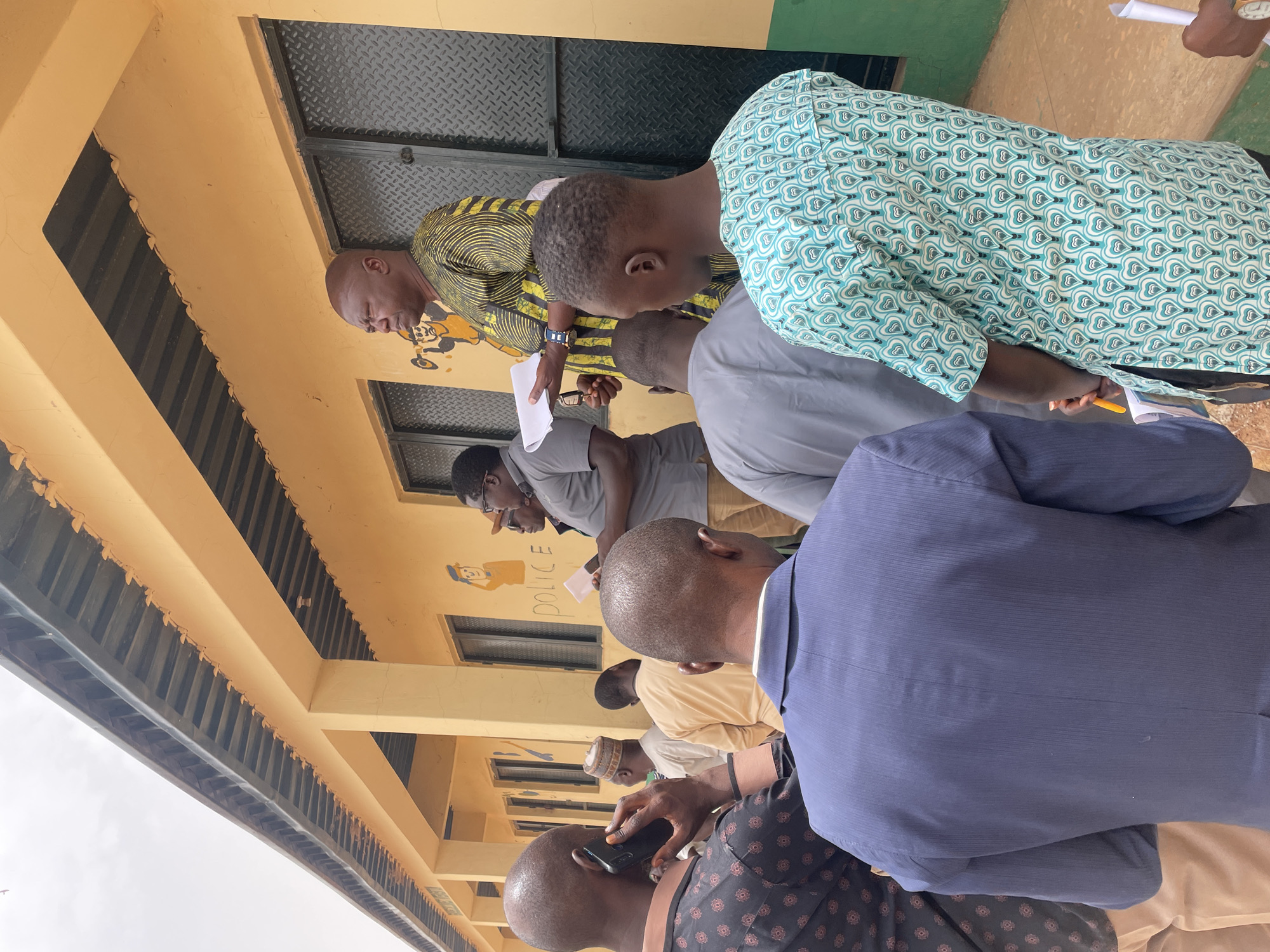The image shows a group of men standing in front of a building in Bukuru, Nigeria. The building is yellow with green trim and has a sign that reads "POLICE" next to a cartoon drawing of a police officer on the wall. The men are dressed in a variety of clothing, including traditional African attire, suits, and casual wear. Several of them are holding papers. One man is talking on a cell phone. The camera angle is from slightly below, looking up at the group.