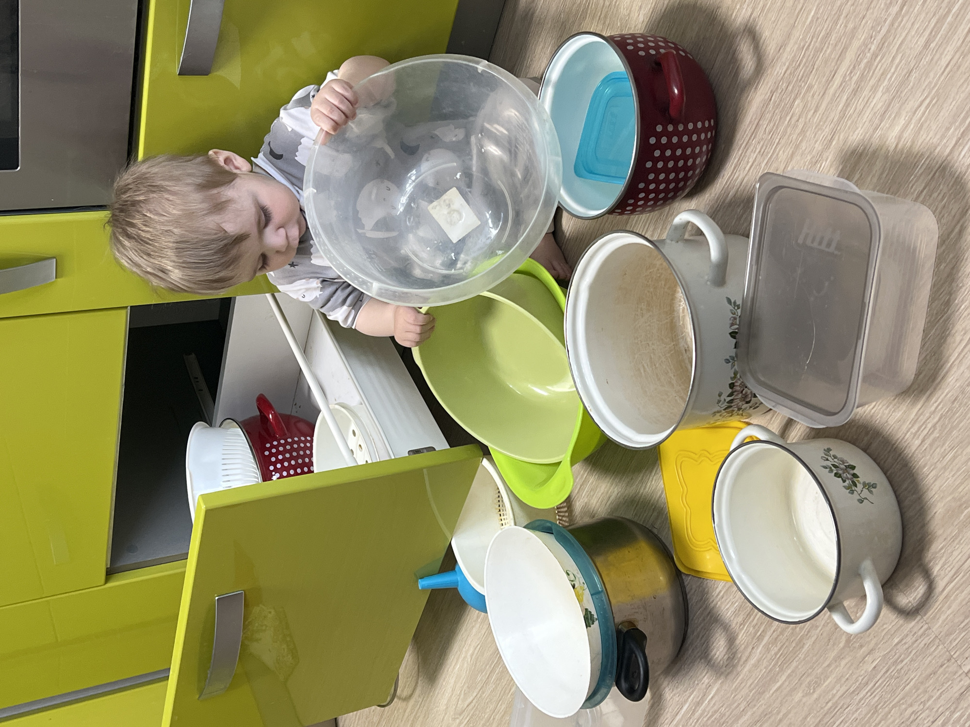 A toddler with blond hair, wearing a gray and white patterned shirt, is reaching into an open lower kitchen cabinet. The cabinet is a bright lime green color. The toddler is holding a clear plastic mixing bowl. A variety of kitchenware items, including pots, pans, bowls, and food containers, are scattered on the floor. The setting appears to be in a residential kitchen in Perm, Russia, as the 'Hutt' food container brand is local.