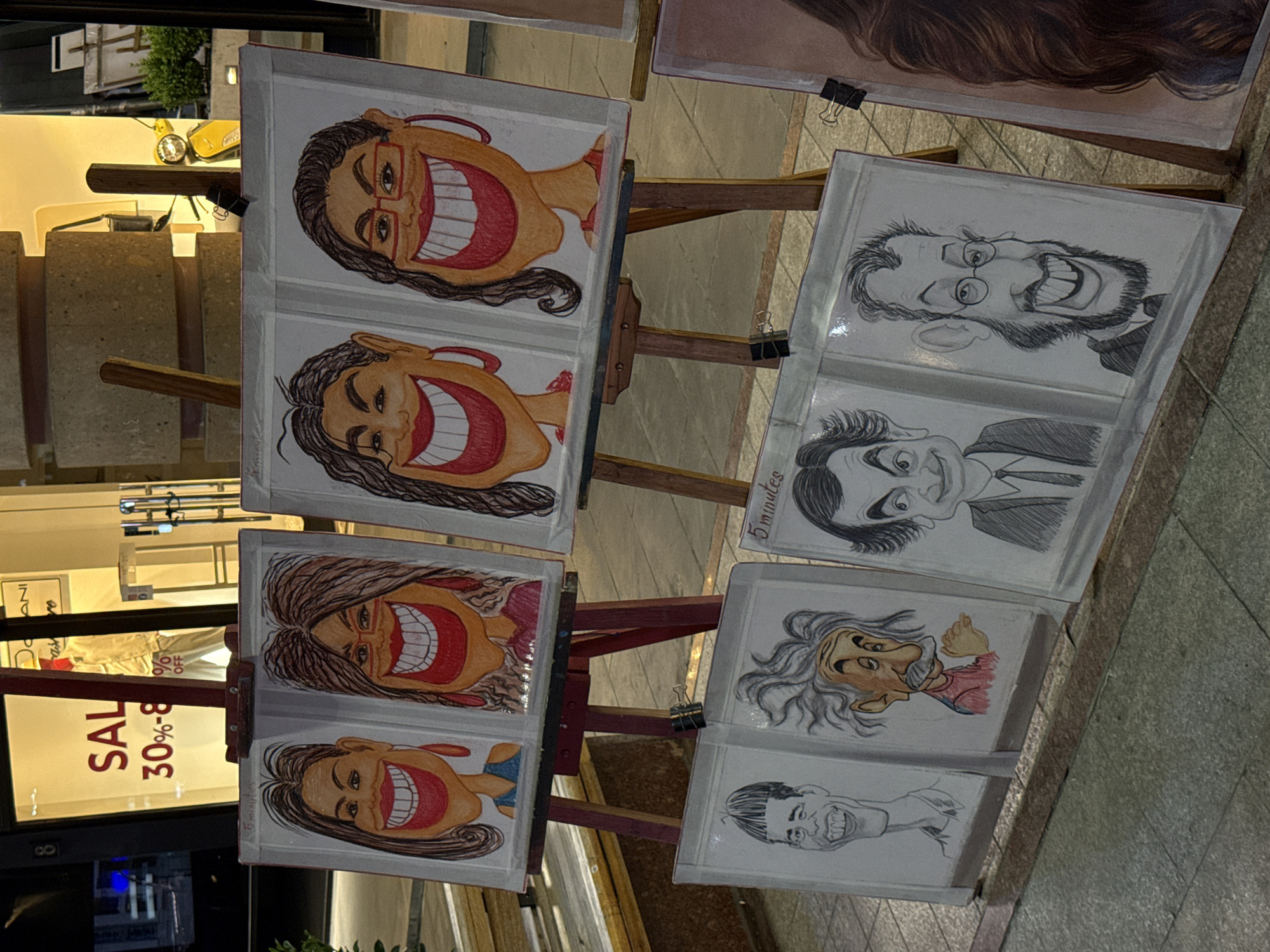 The image captures an outdoor display of caricatures, likely by a street artist, under artificial lighting at night. Several caricatures are presented on wooden easels, held in place by binder clips. The artwork features exaggerated facial features, mostly depicting wide, toothy smiles and open mouths. Some caricatures are rendered in color, with skin tones, red lips, and occasional clothing details, while others are black and white sketches.
On the left side of the frame, a set of four caricatures, primarily of women with long hair and glasses in some cases, are displayed on two easels. These are predominantly in color. On the right, another set of caricatures, mostly black and white, is shown. These include a man with a beard and glasses, another man with slicked-back hair, and a figure resembling Albert Einstein, who appears in color. Below these, there are more black and white caricatures of smiling individuals. One of the white boards explicitly states "5 minutes," possibly indicating the time it takes to draw a caricature.
In the background, illuminated shop windows are visible, casting light on the scene. One prominent window on the left displays a "SALE 30%-8" sign, suggesting a retail environment. The ground appears to be paved, indicating an urban setting like a street or a pedestrian area. The overall atmosphere suggests an evening market or a street performance setting.