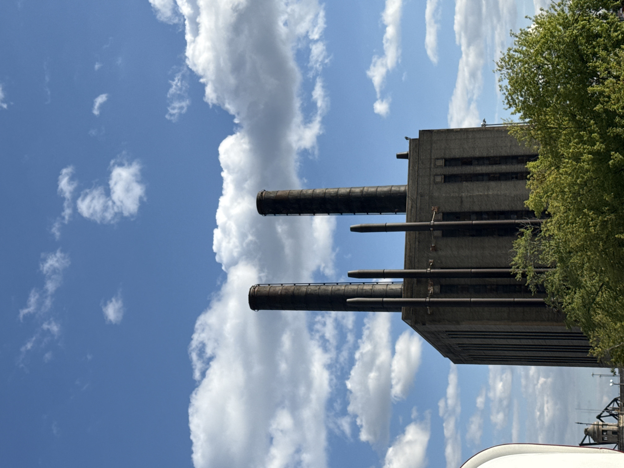 The image shows the Fisk Generating Station in Chicago, Illinois. The station's grey concrete building has multiple smokestacks, and a green tree is visible to the right of the building. The sky is blue with white clouds. Below the structure, there is what appears to be the top of a white structure.