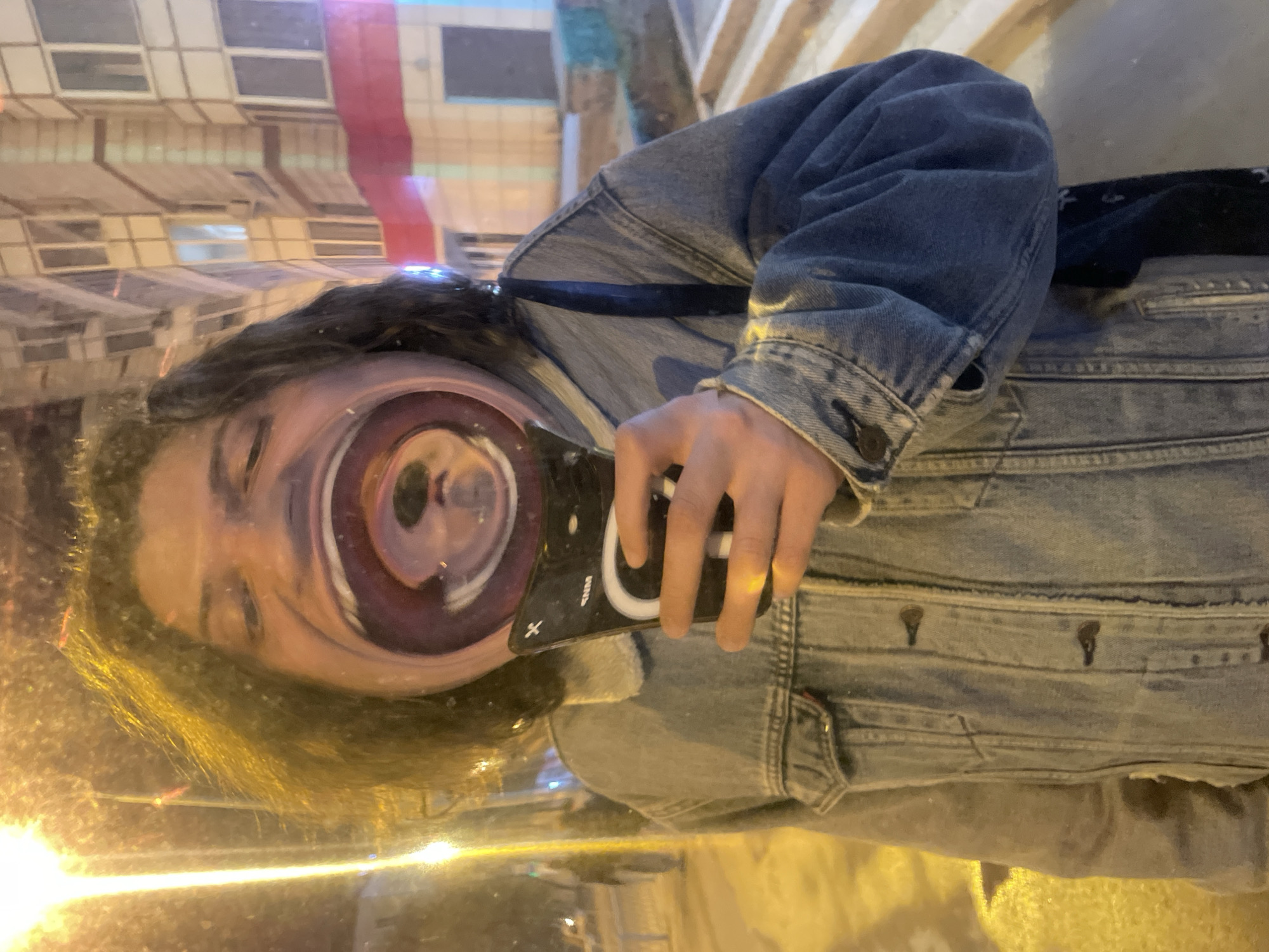A distorted selfie captures an individual in what appears to be an urban environment. The person, wearing a denim jacket, is holding a smartphone with the camera pointed towards their face, creating a warped, funhouse-mirror effect. The background suggests a city street, likely captured in the reflective surface the selfie is being taken in front of. The reflected cityscape hints at the city of Yekaterinburg, Russia, but without clearer details, the exact location cannot be confirmed. The overall impression is that of a playful, self-aware photo in an urban setting.