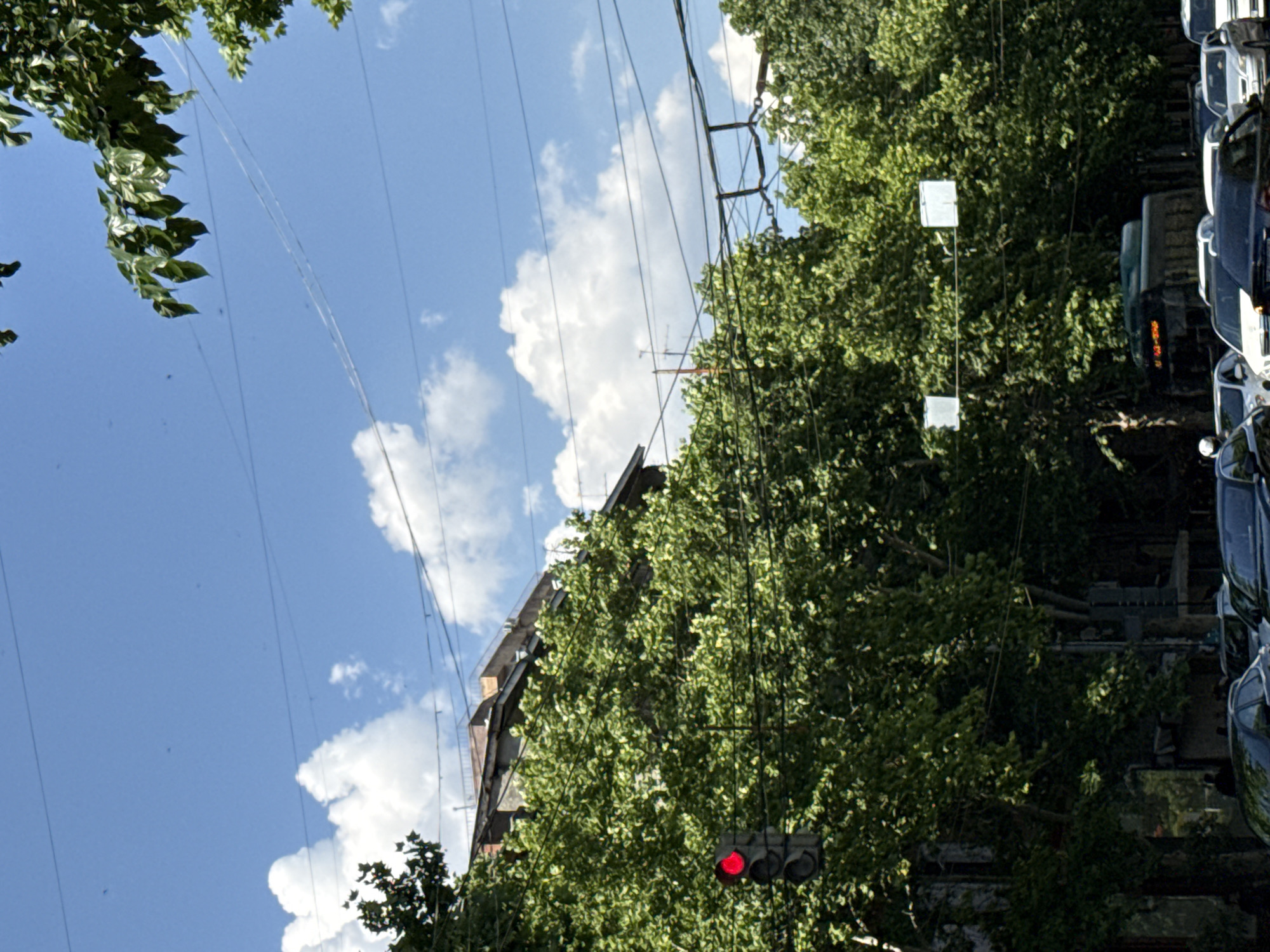 This media file depicts a street scene in Yerevan, Armenia, characterized by a bright, sunny day with scattered white clouds against a clear blue sky. The perspective is slightly tilted, looking upwards towards a cluster of power lines and lush green trees.
Prominently featured are several electrical wires crisscrossing the frame, supported by poles which are partially obscured by the dense foliage. Amongst the trees, a traffic light is visible, showing a single red light, indicating a stop. Adjacent to the trees and power lines, a multi-story building with visible brickwork suggests an urban or semi-urban setting.
To the right side of the image, a line of parked vehicles is visible, appearing to be cars and possibly a truck or bus further back, all reflecting the bright sunlight. The overall atmosphere is one of a typical day in the city, with everyday elements like traffic control, infrastructure, and vehicles present. The time of day is clearly daytime, given the strong sunlight and blue sky. No people are directly visible in the frame. There is no discernible text within the image.