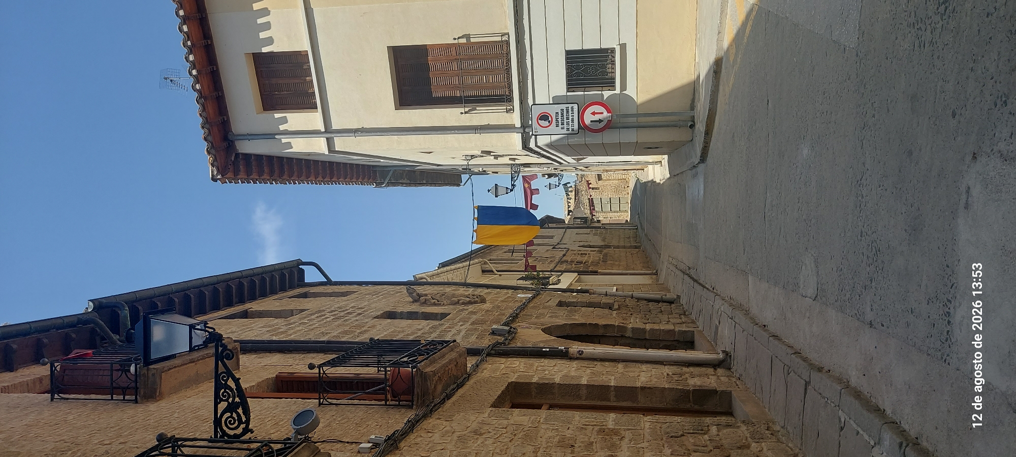 The image captures a narrow, paved street in the historic town of Olite, Spain. The street is flanked by two buildings: on the left, a rustic building constructed with rough-hewn tan stone, featuring wooden doors, balconies with black wrought-iron railings, and decorative black lanterns. On the right, a lighter-colored building with a tiled roof and wooden shutters on its windows and balconies.
Hanging across the street are two decorative banners. The larger banner in the center is split vertically into blue and yellow sections, while smaller red banners with gold trim are visible further down the street. Streetlights are mounted on the building on the left and on a pole on the right.
Traffic signs are visible on the right side of the street. One sign, circular with a red border, depicts a person with a diagonal line through it, and text below reads "RESPETEN EL DESCANSO DE LOS VECINOS DE 22:00h A 9:00h" (Respect the rest of the neighbors from 10 PM to 9 AM). Below this is a circular sign with a red border and a white background, showing two red arrows pointing in opposite directions, indicating traffic flow restrictions.
The pavement of the street is made of rough, grey concrete. The sky above is clear and bright blue, suggesting a sunny day. The image is timestamped "12 de agosto de 2026 13:53".