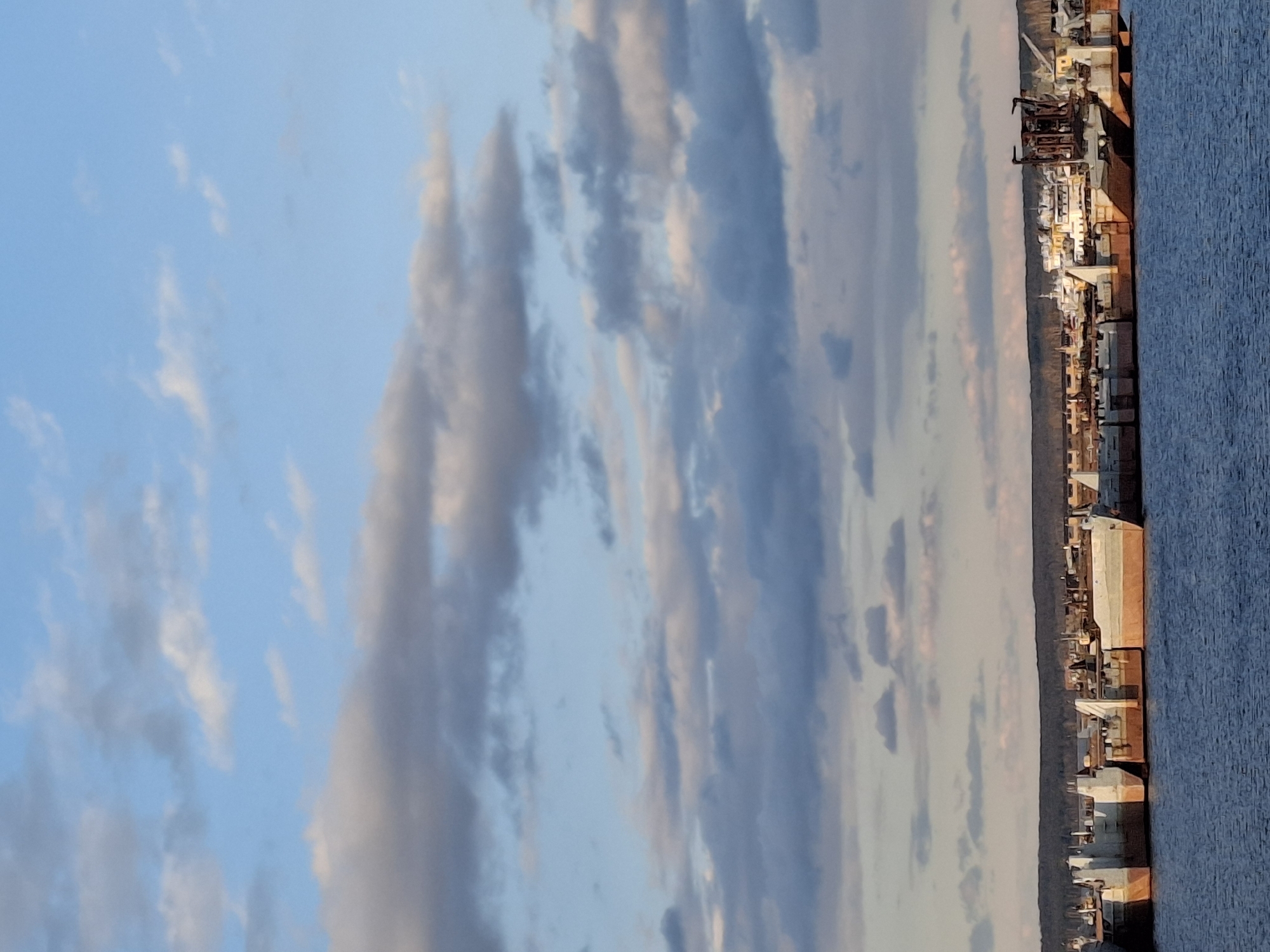 The image, correctly oriented, captures a panoramic view of a large body of water and a distant landmass under a partially cloudy sky. The upper two-thirds of the frame are dominated by a clear, light blue sky scattered with elongated clouds. Some clouds are bright white or light gray, while others display subtle warm hues of pink and orange, indicative of either sunrise or sunset.
Along the horizon, a long strip of land features an array of structures, suggesting an industrial area or port. These buildings and facilities appear to be diverse, with some prominent shapes resembling ships or large machinery docked along the waterfront. The soft, warm light of the setting or rising sun illuminates these structures, casting golden and brownish tones on their surfaces. Behind these foreground structures, a dark, thin line marks the distant land or shoreline.
The lower right portion of the image is filled by a vast expanse of water, a deep blue in color with visible texture, suggesting small ripples or waves. This body of water, given the location context of Chaikovsky, Russia, is likely the Kama River or Votkinsk Reservoir.
No people or explicit activities are visible, and there is no discernible text in the image. The scene conveys a tranquil atmosphere, showcasing a riverside industrial landscape at a transitional time of day.