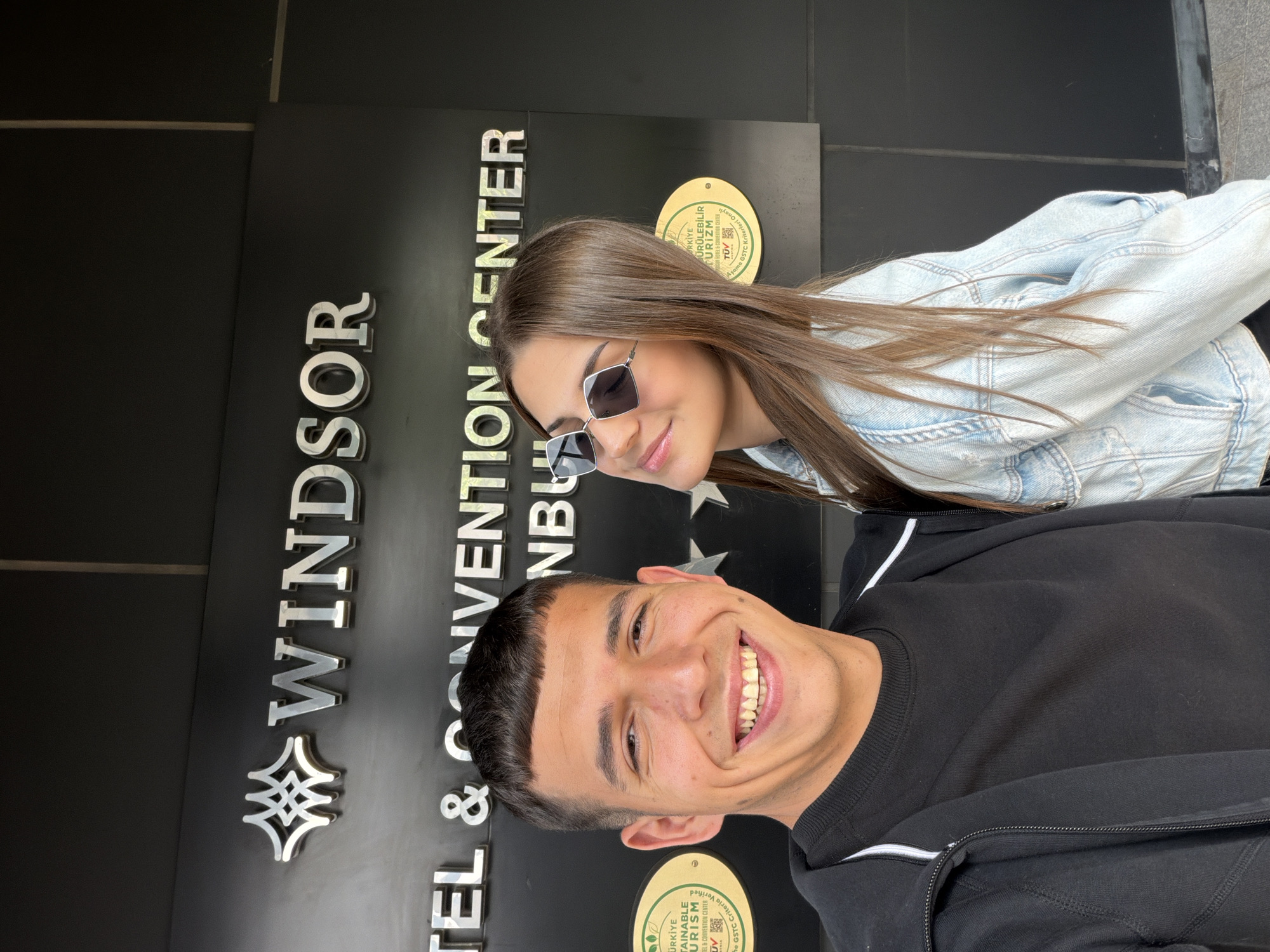 A man and a woman are posing for a photo in front of the Windsor Hotel & Convention Center in Istanbul, Turkey. The man is wearing a black sweater and the woman is wearing a light blue denim jacket and sunglasses. Both are smiling.