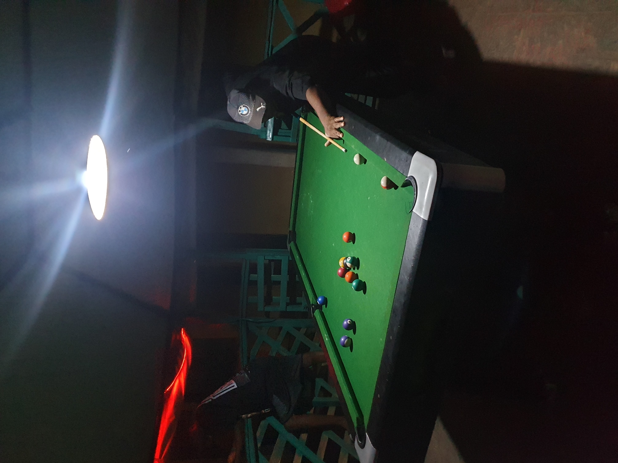 A person wearing a black shirt and a cap with a white logo is playing pool on a green pool table. The pool table has various colored pool balls scattered on its surface. The scene is illuminated by a bright light source, possibly a lamp or spotlight, casting a strong glare in the image. The environment appears to be outdoors, with a patterned fence structure visible. The city of Lokoja, Nigeria, is not explicitly identifiable in the image.