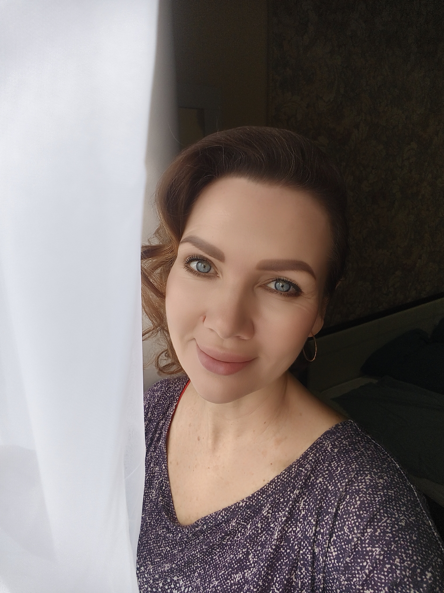 A middle-aged woman with light skin and blue eyes poses for a selfie. She is partly obscured by a white curtain on the left side of the frame. The woman has light brown hair styled in waves, and is wearing a purple and white speckled sweater. She is wearing a small gold hoop earring. The background behind her is a dark textured wall, likely inside a building. The image location is unknown.