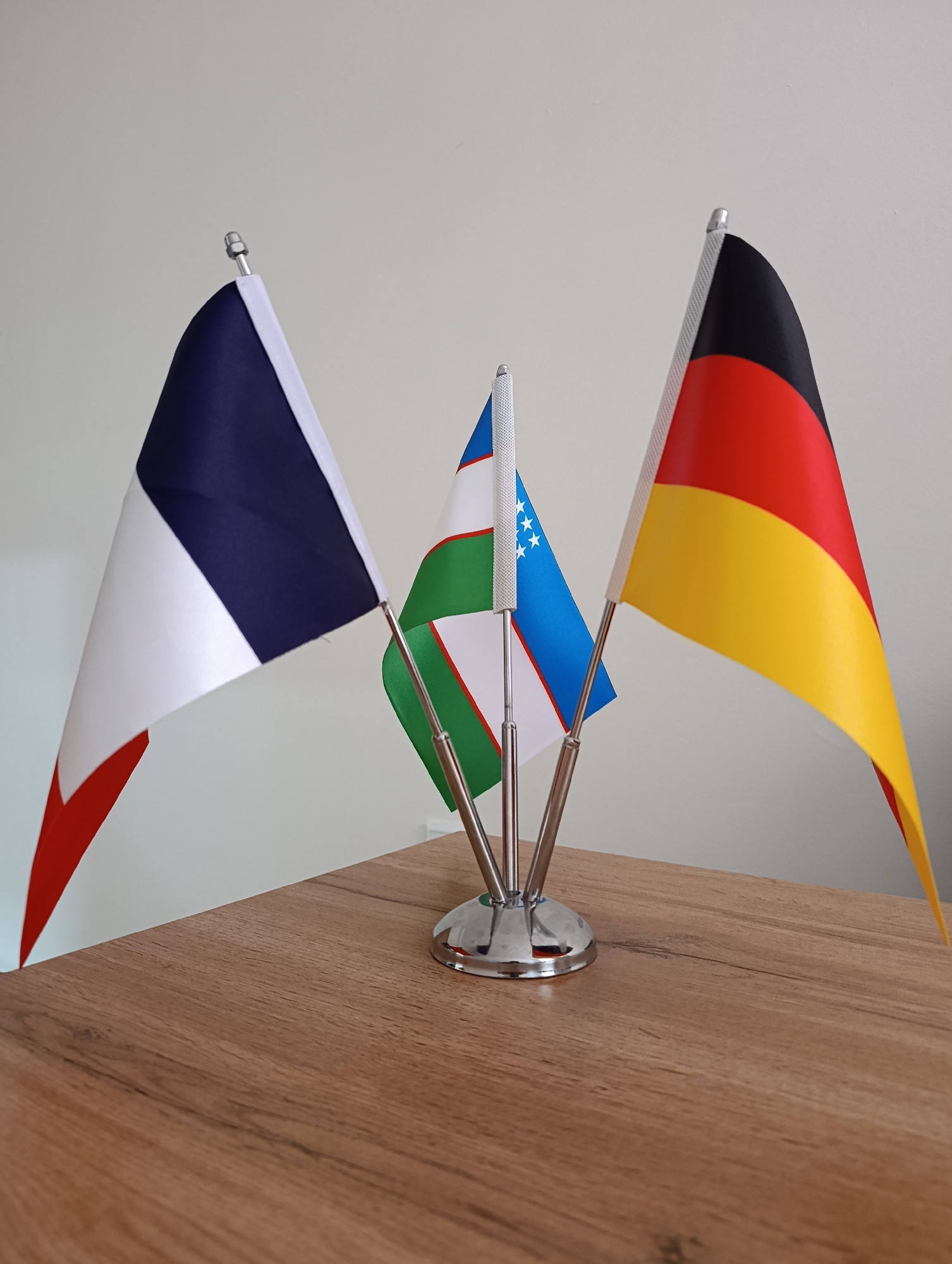 The image displays three small national flags mounted on a chrome stand. From left to right, the flags are those of France, Uzbekistan, and Germany. The French flag has vertical bands of blue, white, and red. The Uzbek flag features horizontal bands of light blue, white, and green, with a crescent moon and twelve stars on the white band. The German flag has horizontal bands of black, red, and yellow. The flags are positioned on a wooden surface against a plain, light-colored background. The scene is indoors, and given the presence of the Uzbek flag, it is possible this image was taken in Uzbekistan, though no specific city or location is identified.