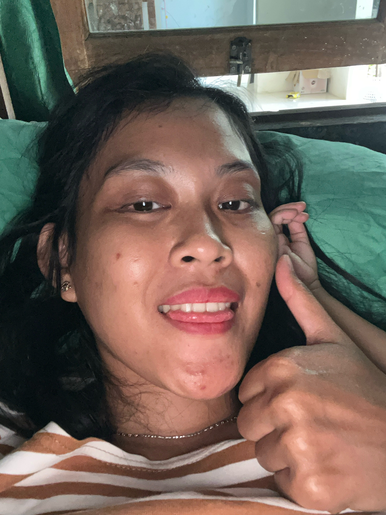 In a close-up, selfie-style shot, a person with dark hair lies down, offering a thumbs-up gesture with their right hand. Their mouth is slightly open, with their tongue peeking out, and their lips are glossed. A delicate silver necklace adorns their neck, resting on a striped brown and white shirt. The background reveals a green fabric, possibly a curtain or blanket, and a wooden frame with a latch, suggesting an indoor setting. The lighting appears to be natural and diffused. There is no specific indication of the location being in Kecamatan Pati, Indonesia, beyond the user's prompt.