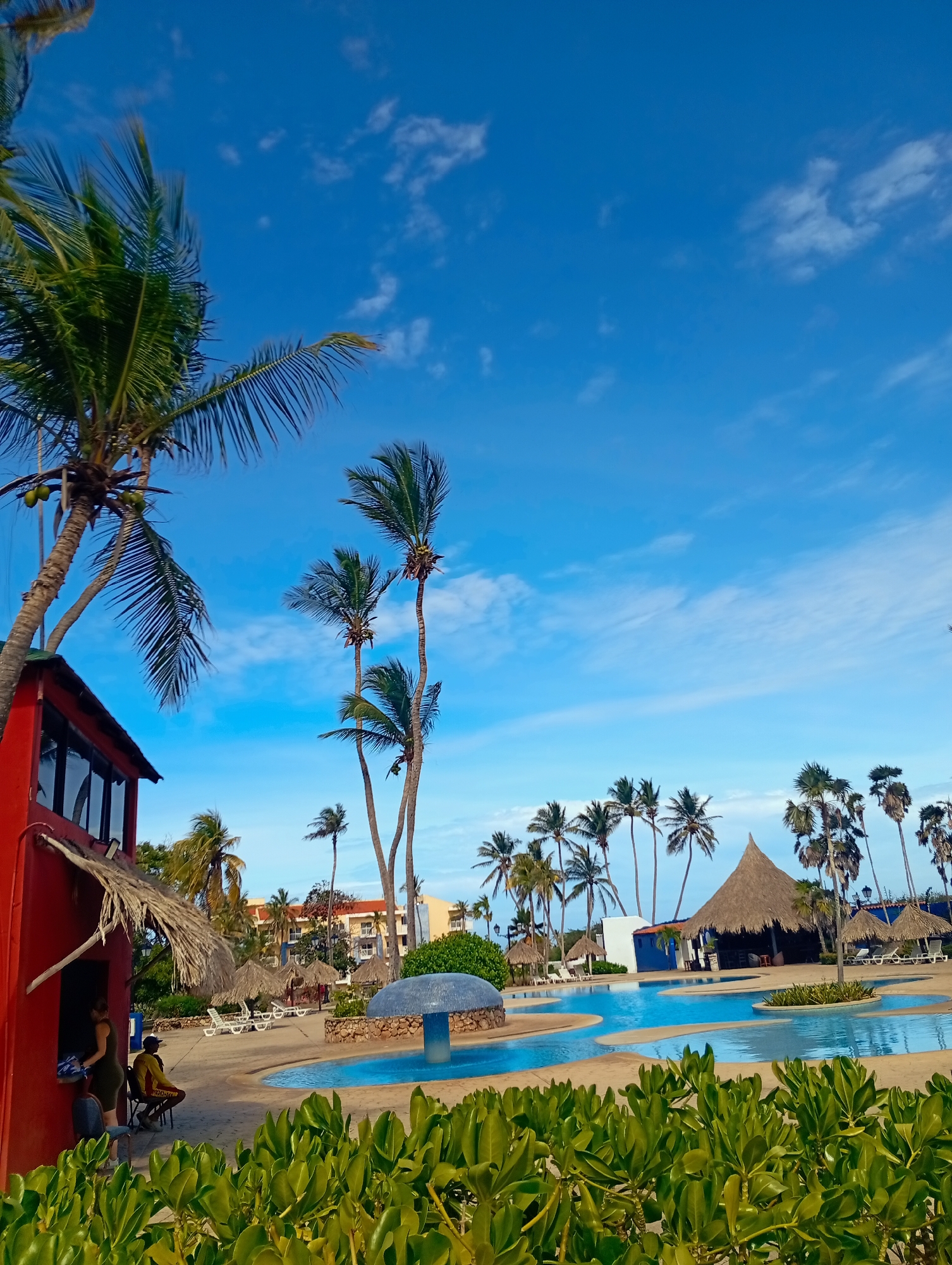 The image depicts a sunny outdoor scene, likely at a resort in Playa El Agua, Venezuela. In the foreground, lush green shrubbery lines the bottom of the frame. A multi-tiered pool area dominates the center. In the middle ground, palm trees are scattered throughout, including several tall, slender palms. A thatched-roof structure stands adjacent to the pool. At the left edge of the frame is the corner of a red building with a thatched roof extension. Two people are visible near the building. The sky is blue with scattered white clouds. A building is visible in the mid-ground behind the palms and pools.