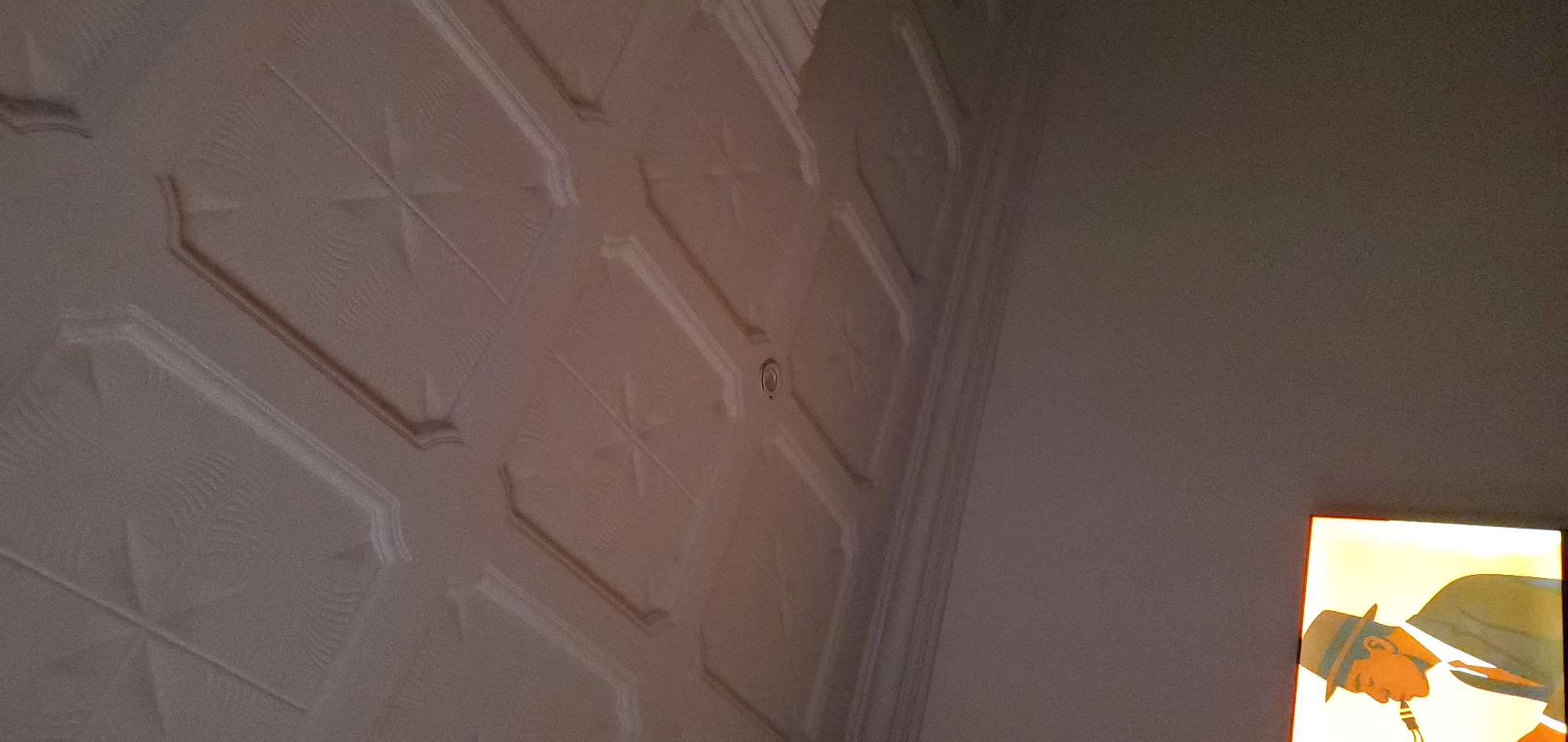 The image shows a portion of a decorative ceiling with recessed panels and a small, circular object (likely a light fixture or smoke detector) in one panel. To the right of the ceiling is a plain wall, and in the lower right corner, a partially visible framed image of a man with a hat is seen. The location is indoors. The city is Unknown, Unknown.