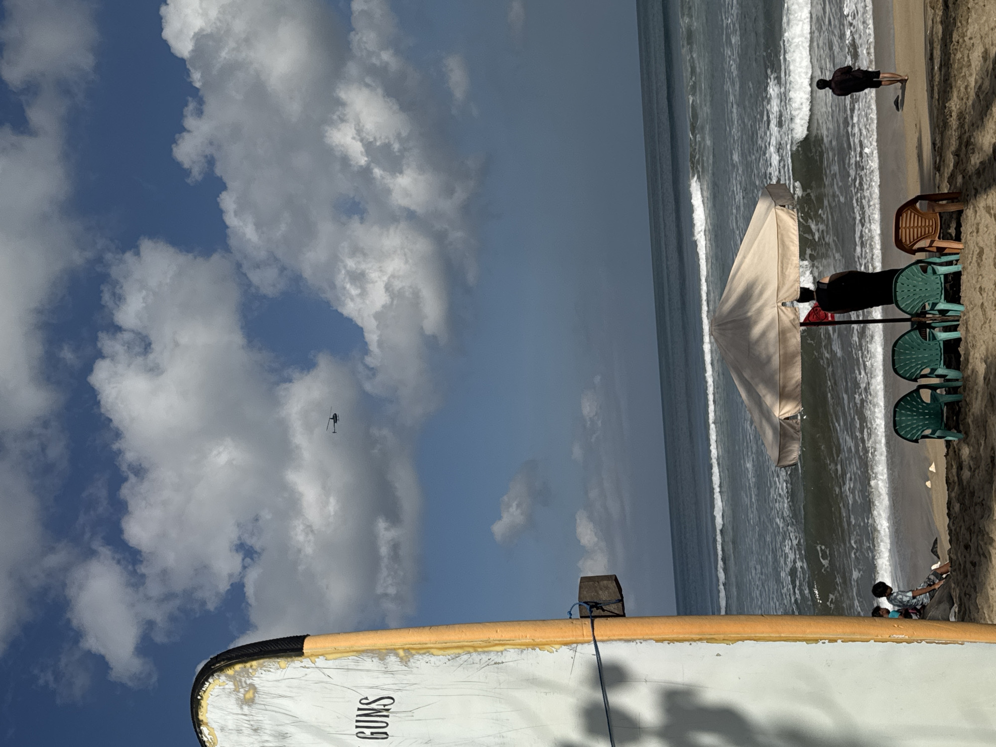 A wide shot captures a sunlit beach in Kuta, Indonesia, with a clear blue sky dotted with fluffy white clouds. In the distance, a helicopter is visible against the sky. In the foreground, a white surfboard with orange trim stands upright on the sand. Several plastic chairs, including two green and one orange, are arranged on the beach. A large, light-colored umbrella provides shade over a sandy area. Two people are seen near the water, one walking away from the camera and another standing under the umbrella. The ocean waves are breaking on the shore. The scene is bathed in bright sunlight, casting shadows on the sand.