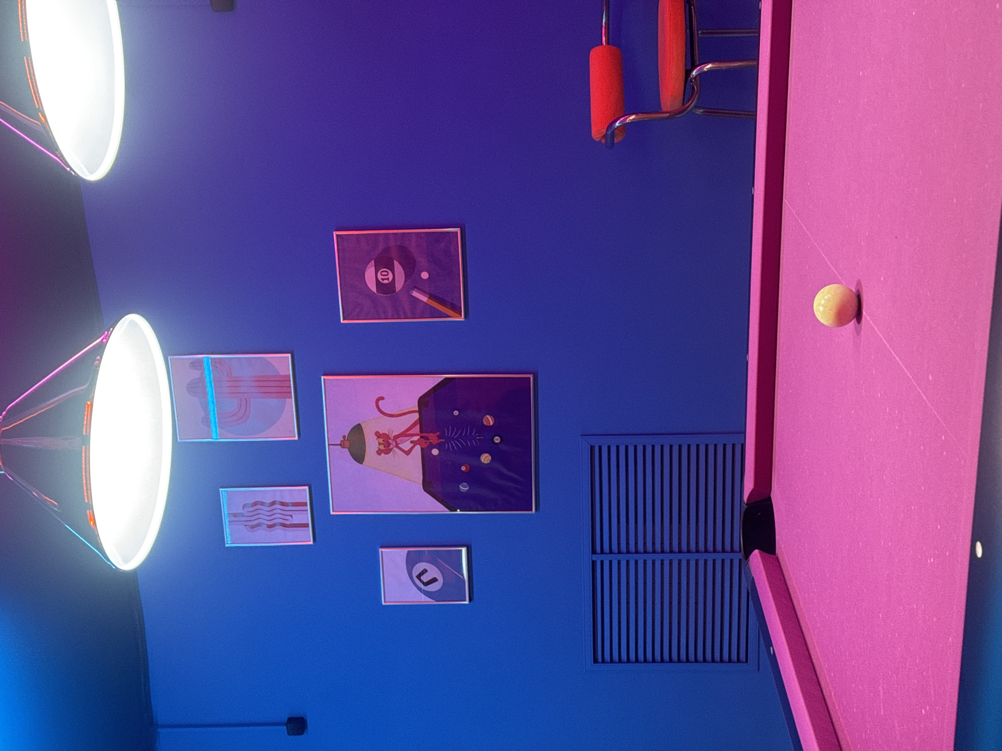 This image depicts a vibrant and colorful indoor recreational space, likely in Krasnoyarsk, Russia. The scene is dominated by a deep blue wall on the left, adorned with several framed artworks, and a striking pink pool table on the right.
The pool table is a central feature, showing a light-colored cue ball resting on its pink felt near a black pocket. Partially visible on the far right, above the pool table, are parts of red-padded chairs or stools with chrome frames, suggesting a seating area. Two large, white circular pendant lamps hang from above on the upper left, casting bright artificial light and illuminating the space.
The blue wall is decorated with an eclectic collection of framed prints. Visible artworks include: an illustration of an 8-ball with a cue stick; another depicting a 7-ball; a larger piece featuring a character resembling the Pink Panther with a futuristic or space-themed pool scene; and two more abstract pieces with blue lines and wavy patterns. A dark, louvered ventilation grate is also visible on the blue wall, beneath the chairs.
The artificial lighting suggests an indoor setting, likely in the evening or at night. The contrasting colors of the deep blue wall and the bright pink pool table create a modern and energetic atmosphere. No people are visible, but the setup implies a setting for playing pool.