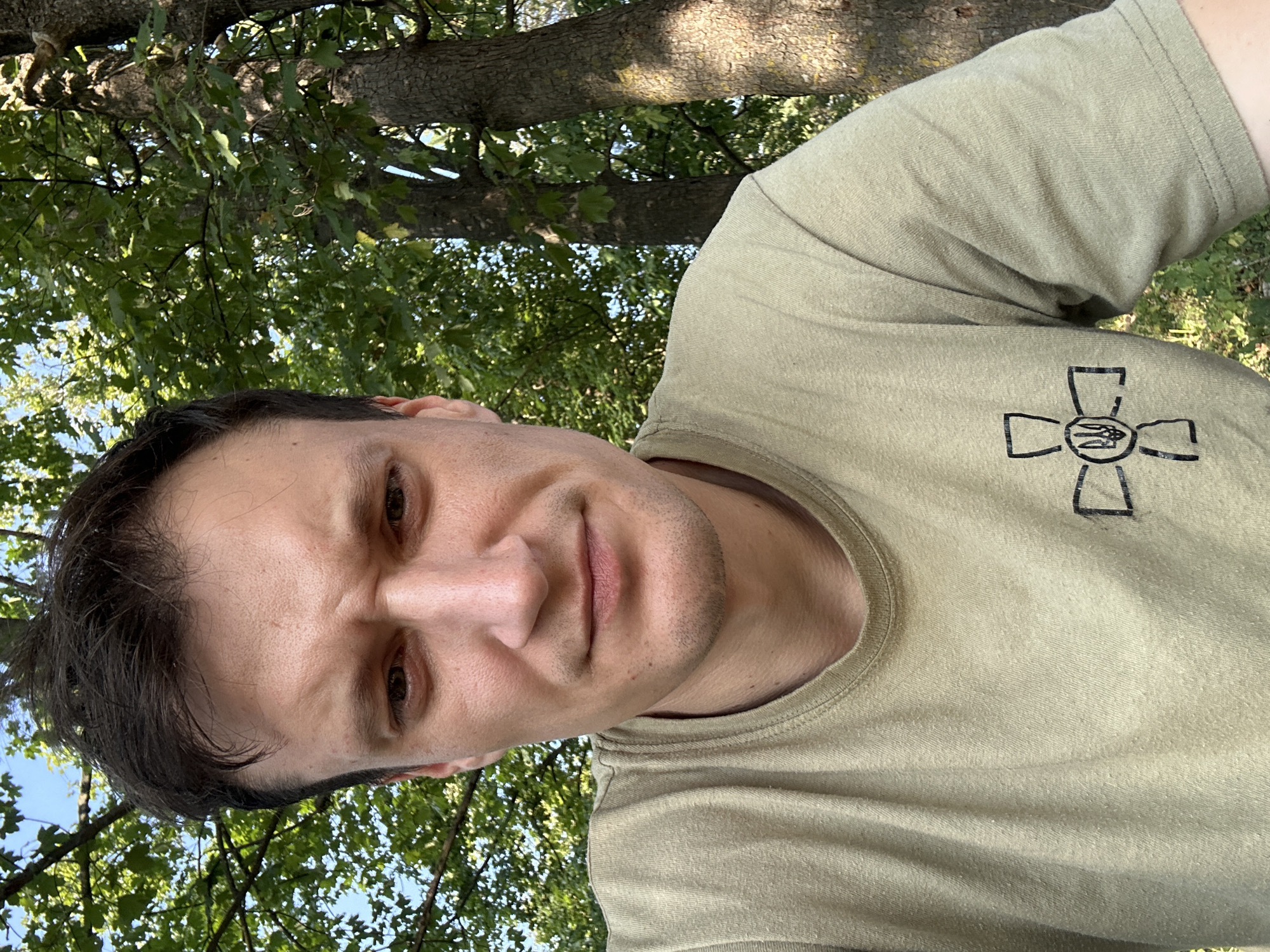 The image is a close-up, selfie-style portrait of a man wearing a light olive-green t-shirt with a black emblem on the left chest. The emblem is a stylized cross with a trident in the center. The man has dark hair, a light complexion, and is looking directly at the camera with a slightly furrowed brow. He appears to be outdoors, with green trees and foliage visible in the background, as well as a blue sky. The lighting suggests it is daytime. The image composition focuses on the man's face and upper body. There is no identifiable location within the city of Makiv, Ukraine, visible in the image.