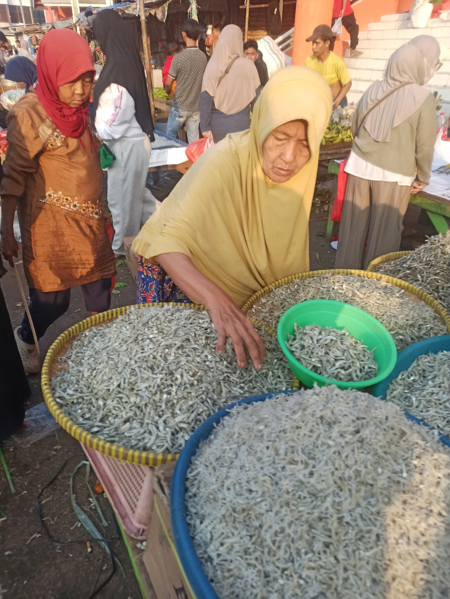An outdoor market scene in Cikarang, Indonesia, shows several people and large quantities of dried fish. In the foreground, a woman wearing a yellow headscarf and a floral patterned skirt is reaching into a large, shallow woven basket filled with small, dried fish. Beside this basket is a bright green plastic bowl also containing dried fish. Behind her, another woman in a brown tunic and red headscarf stands looking down. To her left and slightly behind is a woman wearing a black headscarf and white jacket. Other individuals are visible in the background, including a man wearing a yellow shirt and a baseball cap, and others obscured by distance and activity. The ground appears to be a dirt or paved surface. Several woven baskets and plastic containers filled with fish are present, suggesting a fish market stall. The overall lighting indicates it is daytime, with sunlight casting shadows.