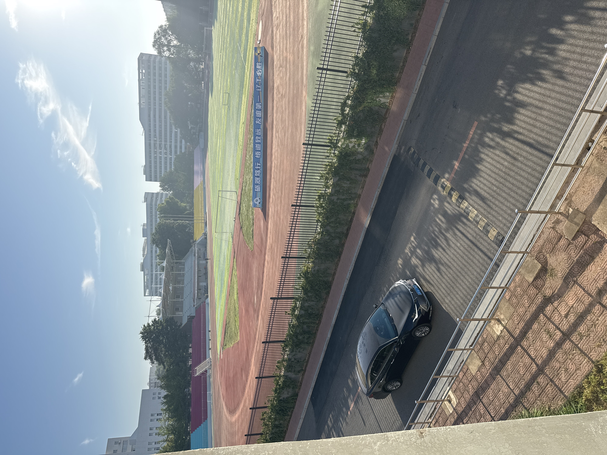 The image depicts a view of a running track and sports field from an elevated position, likely a building. The running track has red and blue sections, with a green field in the center. Bleachers with red and blue seating are visible on the left side of the track. Buildings, some with multiple stories and modern architecture, are situated in the background, surrounded by trees. The sky is clear with a few wispy clouds, and the sun appears to be shining, casting shadows on the scene.
In the foreground, a black sedan is parked on an asphalt road that runs alongside the track, separated by a black metal fence topped with ivy. The road has yellow and black speed bumps. Shadows from nearby trees are cast across the road and a paved walkway with a patterned surface in the bottom right of the frame. A light-colored concrete structure forms the immediate foreground on the left.
The scene appears to be a sports facility within a university or educational institution, given the architecture of the buildings and the presence of a running track. The location is identified as Zhongtun, China. The overall atmosphere is serene and suggests it is either early morning or late afternoon due to the quality of light and the long shadows. No people are clearly visible or engaged in any activity.
