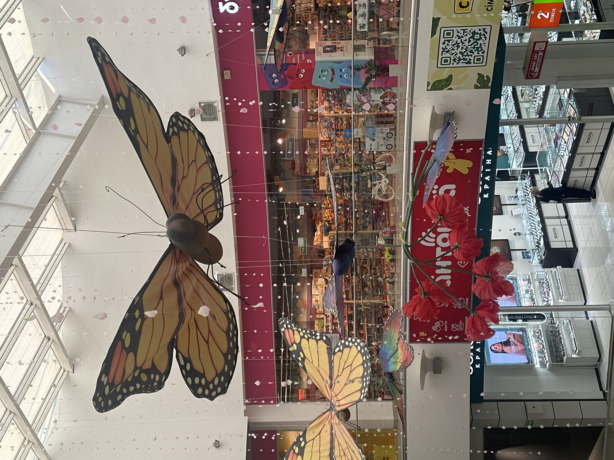 The image shows a retail environment, likely a shopping mall, with decorative elements hanging from the ceiling. Large, artificial monarch butterflies are suspended by wires, with smaller, colorful butterflies also present. Pink and white flower petals are scattered and suspended on thin threads throughout the space, creating a festive or seasonal atmosphere. Below these decorations, multiple storefronts are visible, including a jewelry store with displays of watches and necklaces. One storefront features a large screen displaying a woman. A QR code is visible on a light green banner advertising "SPORT LIPE". A person is standing inside the jewelry store, interacting with a display. The overall impression is a brightly lit indoor commercial space.