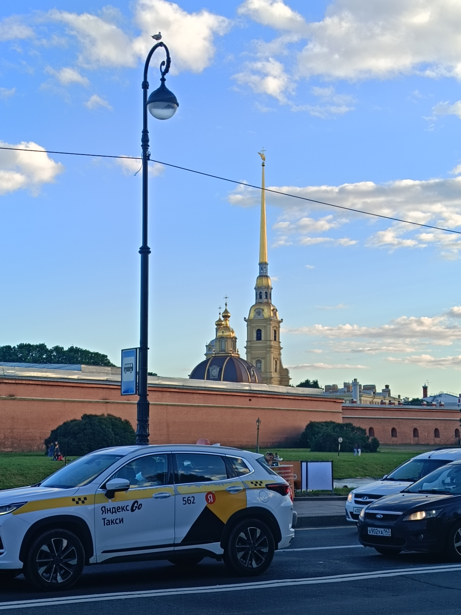 This is a medium-distance shot of a street scene in Saint Petersburg, Russia, featuring a section of the Peter and Paul Fortress wall and its iconic spire, with several cars in the foreground. The sky is blue with scattered white clouds. A tall, black lamppost with an ornate lantern stands on the left, topped by a seagull. A bus stop sign is attached to the lamppost.
In the middle ground, a red brick wall of the Peter and Paul Fortress stretches across the frame, with green trees and bushes planted along its base. Behind the wall, several buildings are visible, most notably the golden domes and the tall, gilded spire of the Peter and Paul Cathedral, a prominent landmark in Saint Petersburg.
In the foreground, a white Yandex Go taxi is parked on the right side of the street. It has yellow and black branding, including the Yandex Go logo and the word "Такси" (Taxi). A dark-colored car is behind it, and another white car is partially visible to the left of the Yandex Go taxi. The road surface shows white lane markings. People can be seen walking and sitting on the grass in the background, near the fortress wall.