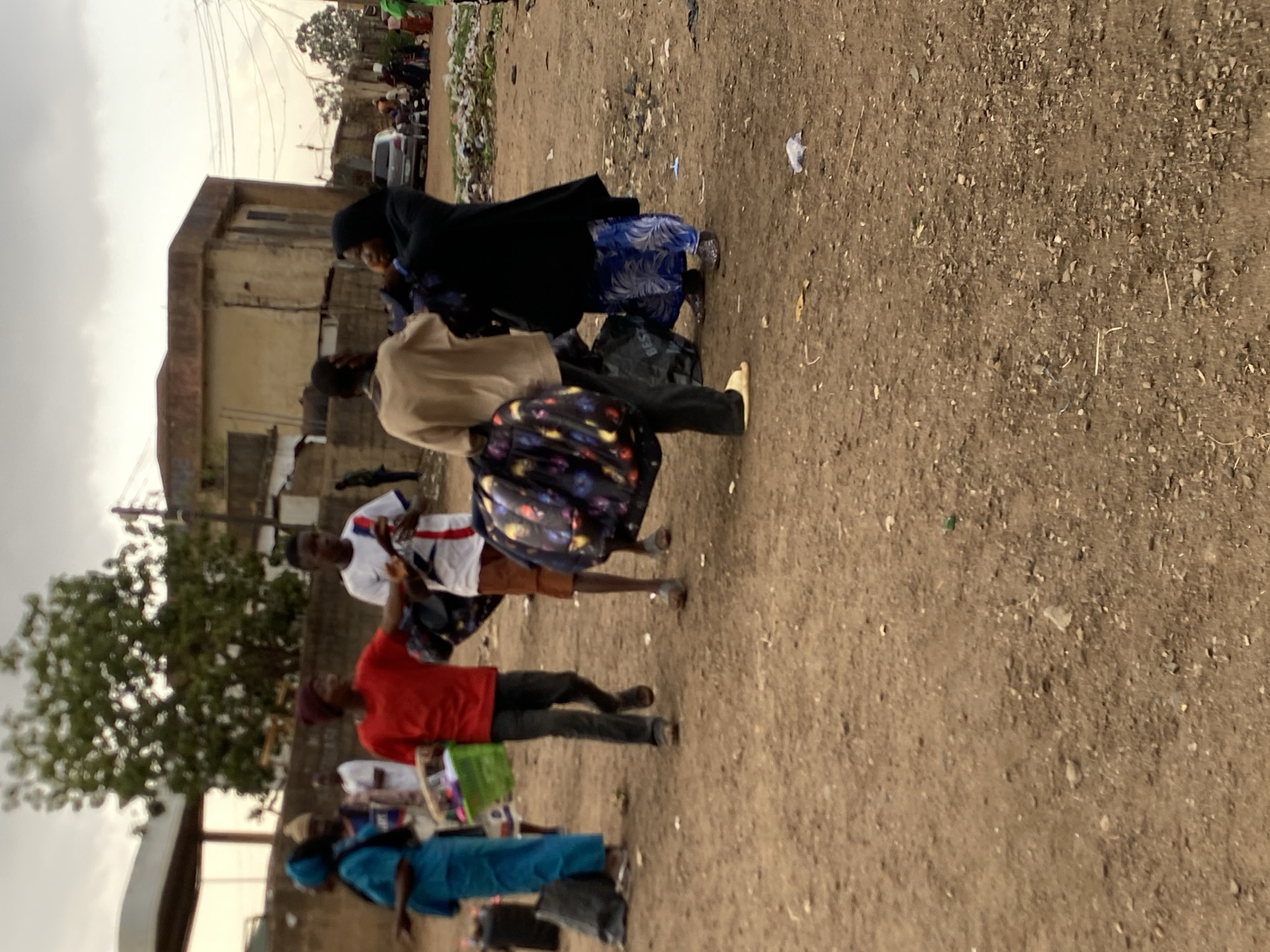 The image depicts a street scene in Bukuru, Nigeria. A group of people are walking along a dirt road. One woman is wearing a black hijab and a blue patterned dress. Another individual is wearing a tan top and a colorful patterned skirt. A person in a red shirt and black pants is also visible. The ground is dirt and scattered with debris. There is a building in the background and some vegetation on the left side. A car can be seen further down the road. The sky appears overcast.