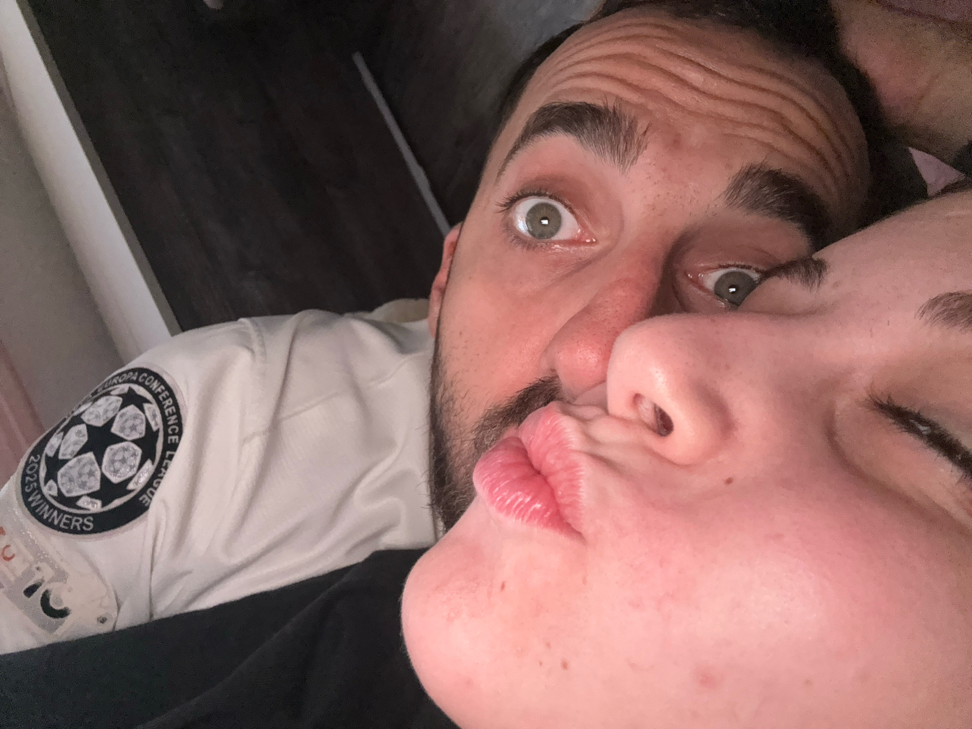 The image shows a close-up, selfie-style photograph of two people. One person, a male with a beard and wide eyes, is looking directly at the camera. The other person, a female, is leaning in to kiss the male, with her lips puckered and touching his cheek. The male is wearing a white jacket with a circular patch on the sleeve that reads "EUROPA CONFERENCE LEAGUE 2023 WINNERS". The background is blurred and appears to be an indoor setting, possibly a room with wooden furniture. There is no identifiable information in the image that suggests the location is in Bulgarovo, Bulgaria.