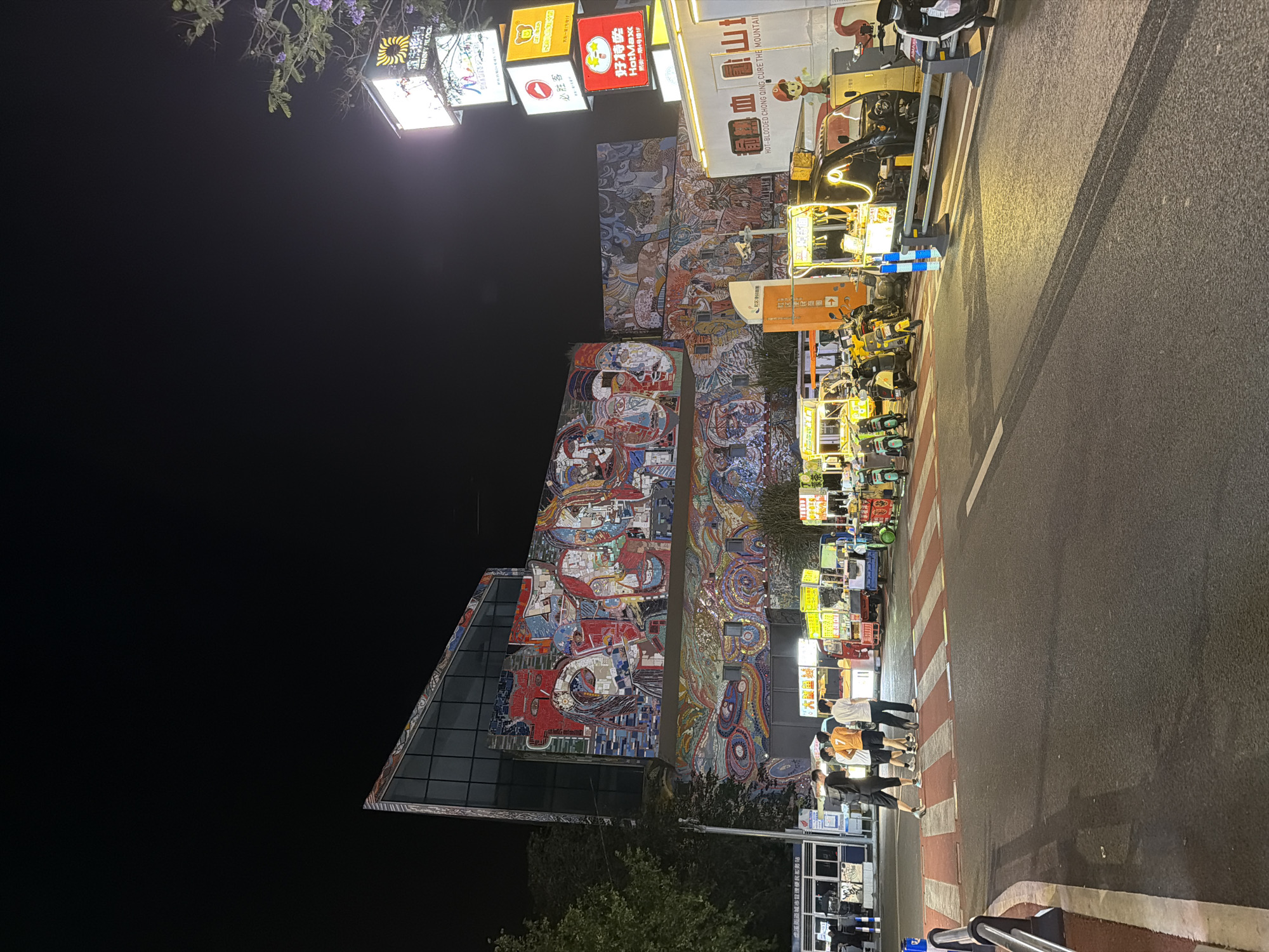 The image depicts a vibrant street scene in Chongqing High-tech Zone, China, at night. The focal point is a tall building adorned with a colorful mosaic mural. Brightly lit signage in Chinese and English, advertising businesses such as "HotMaxx" and "Pizza Hut," are mounted near the top of the building. A row of electric scooters are parked along the street, next to food vendors. Several people are standing on a crosswalk, waiting to cross the street. The asphalt street is visible, partially illuminated by streetlights and lights from the businesses. The sky above is dark.