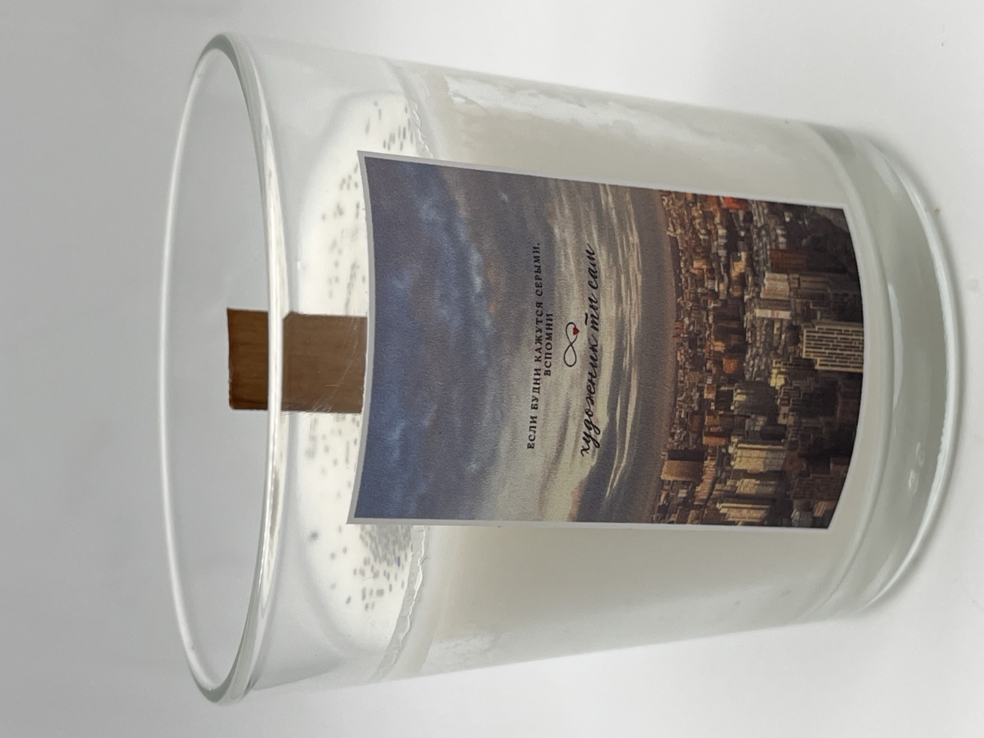 A still life product photograph captures a clear glass candle jar, presented horizontally on a plain white background. The jar contains solid white wax, and a flat, rectangular wooden wick is visible, rising from the center of the wax. The candle appears unlit.
A rectangular label is affixed to the side of the glass jar. This label features a panoramic image of a cityscape, rich in detail, with numerous buildings and skyscrapers. The scene depicts what appears to be either dusk or dawn, with a dramatic, cloudy sky transitioning from lighter hues to darker, potentially stormy tones. The cityscape is illuminated, suggesting lights from the buildings. Due to the candle's orientation, the cityscape on the label is shown rotated ninety degrees to the left.
The label also displays text in Cyrillic (Russian). Part of the text reads: "ЕСЛИ БУДНИ КАЖУТСЯ СЕРЫМИ, ВСПОМНИ" (If weekdays seem gray, remember). Below this, a stylized phrase, possibly a signature or a brand tagline, states: "художник ты сам" (you are the artist yourself). An infinity symbol intricately combined with a small heart is also visible on the label. This Russian text is consistent with the provided location context of Perm, Russia. Inside the jar, above the wax, the glass surface shows small, white, irregular speckles, possibly remnants of the pouring process or reflections. The plain white background emphasizes the product, which embodies a theme of inspiration and creativity.