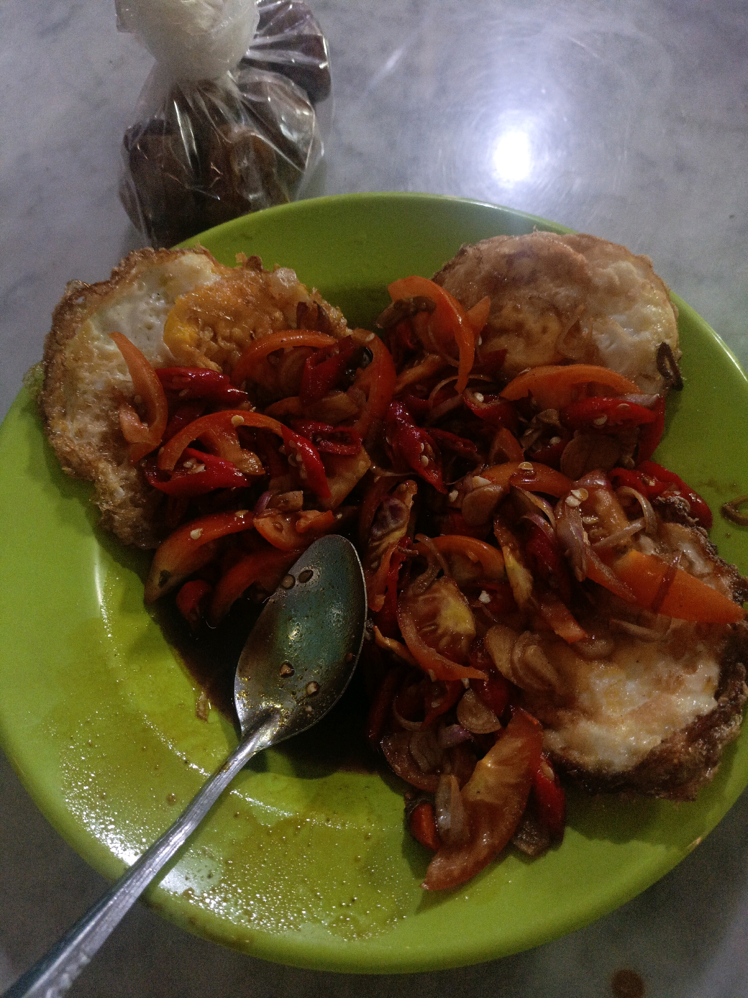 A close-up, overhead shot shows a bright green plate filled with two fried eggs topped with a mixture of sliced red chilies, tomatoes, and onions in a dark sauce. A silver spoon rests on the plate. In the blurred background, a white plastic bag contains small, brown, rounded objects, possibly food items. The setting appears to be a table with a reflective surface, possibly in Undaan, Indonesia.
