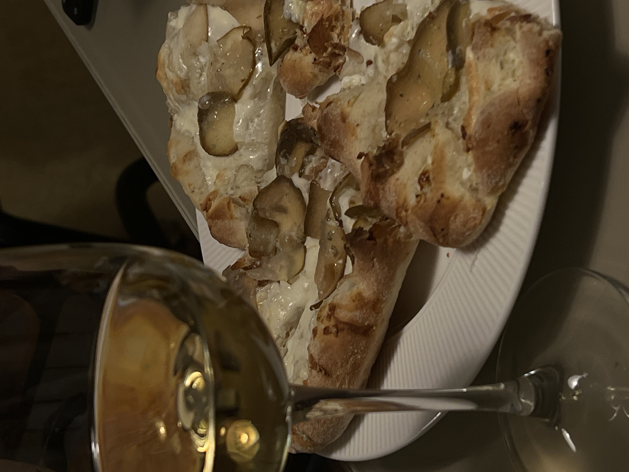 The image depicts a partially eaten white pizza with mushrooms on a white plate. A wine glass filled with a light-colored liquid is visible in the foreground. Another glass is visible in the background. The location is unknown, so I can not confirm if this is Khimki, Russia.