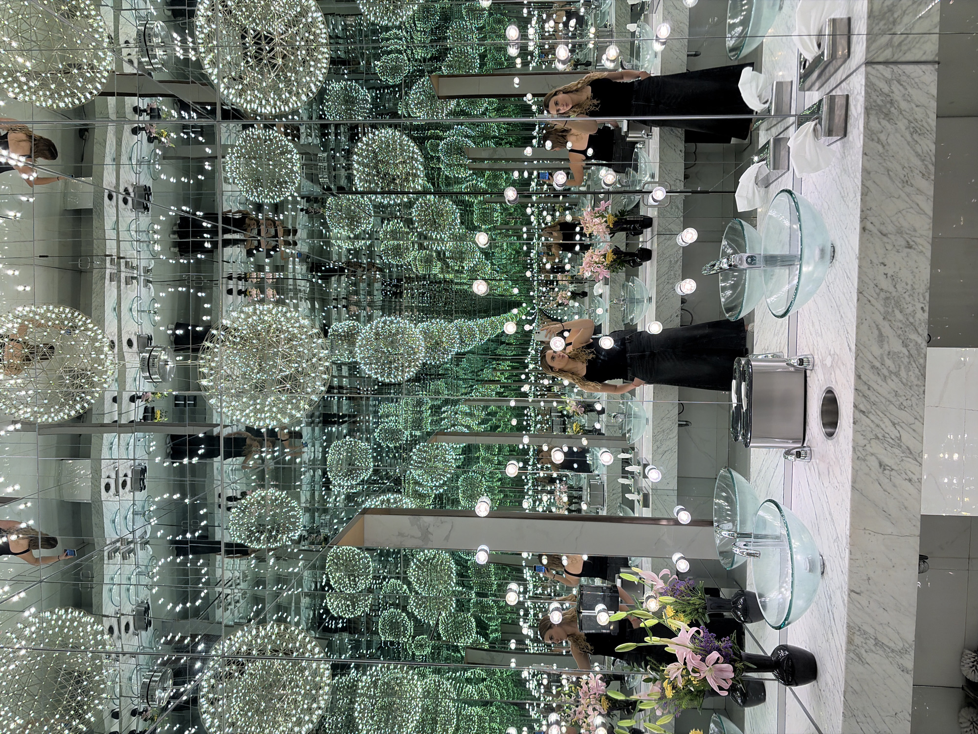 The image captures an infinity mirror effect within what appears to be a restroom, decorated with numerous sphere-shaped light fixtures hanging from the ceiling. The restroom countertop, which is white marble, features a clear glass bowl sink, a silver faucet, and a cylindrical stainless-steel paper towel dispenser. A black vase containing pink and yellow flowers adds a touch of color to the scene. A woman with light hair, wearing a black top, is present in the mirror's reflection, holding a smartphone. The restrooms location is unknown.