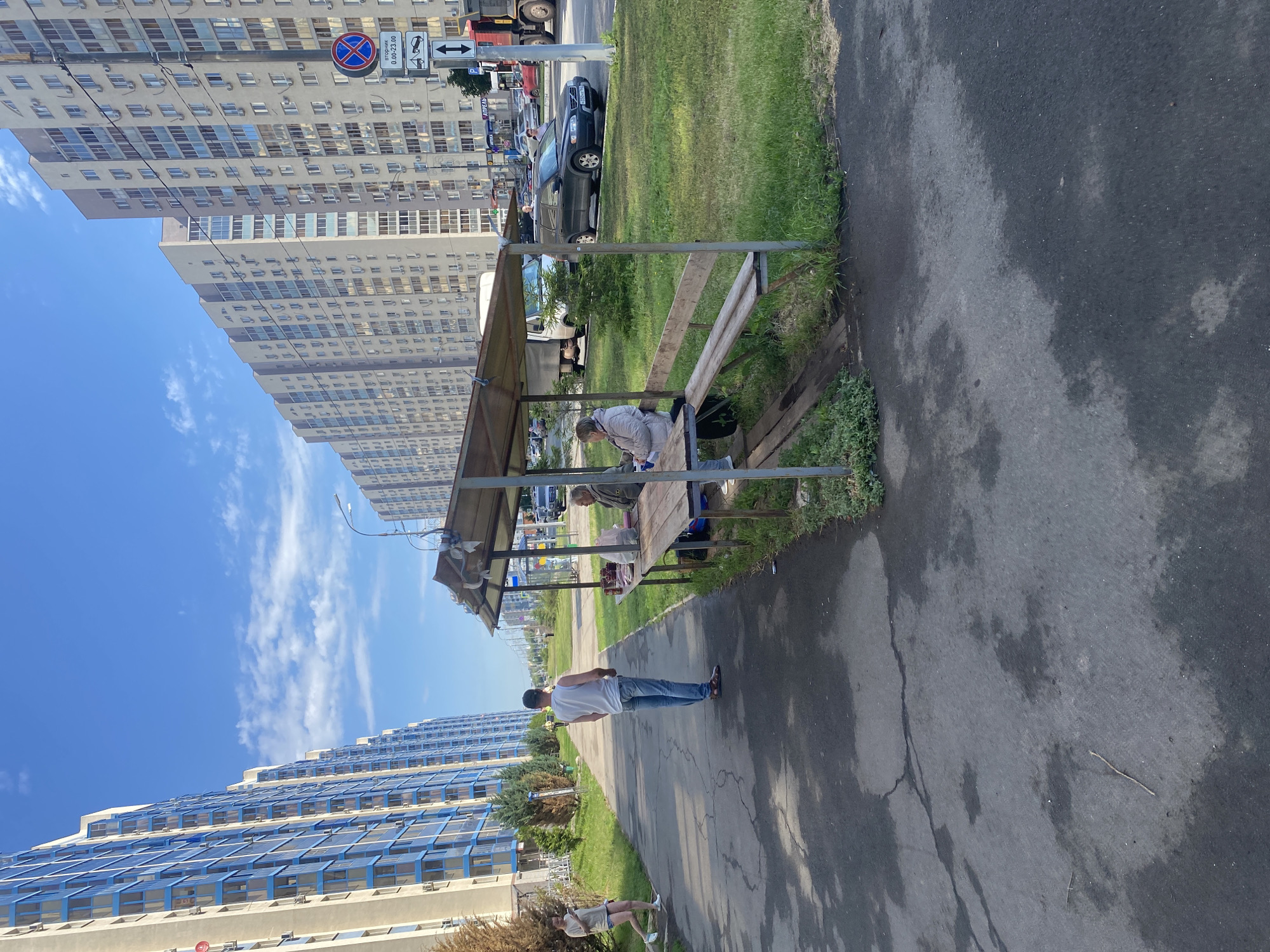 The image depicts a street scene in Rozhdestveno, Russia, with a row of tall, modern apartment buildings on the right and left sides of the frame. In the foreground, a paved walkway runs alongside a grassy area. A makeshift covered seating area with wooden benches and a table is situated on the right side of the walkway. Two elderly individuals are seated at the table, and a bag of groceries rests on the table. A young man in a white t-shirt and blue jeans stands on the walkway, facing away from the camera. Further down the walkway, a young woman in shorts and a t-shirt is walking. In the background, several cars are parked along the street, and traffic is visible, including a truck and a white van. A street lamp is positioned above the covered seating area. The sky is clear blue with scattered white clouds.