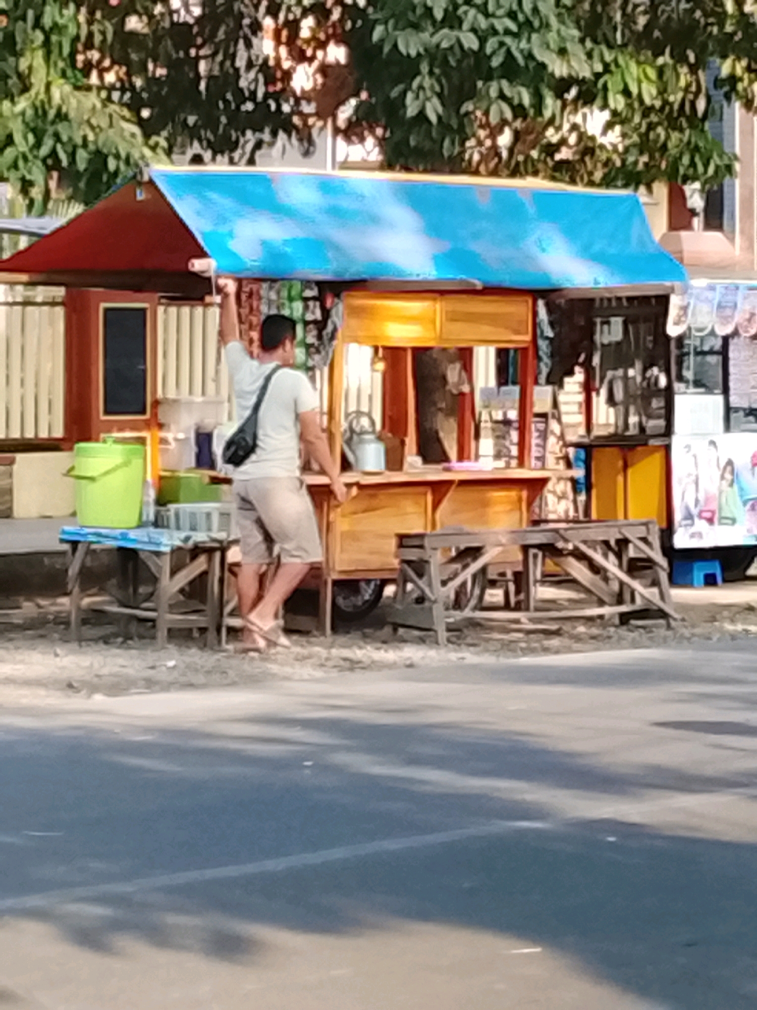 In Slawi, Indonesia, a man is standing in front of a food stall, painting its roof. The stall is made of wood and painted red and blue. The man, wearing a white shirt and shorts, is holding a paint roller and is mid-action. Next to the stall, there are two wooden tables with various items on them, including a green bucket and some containers. In the background, there are trees and a building. The stall appears to be located on the side of a road, with a portion of the asphalt visible in the foreground.